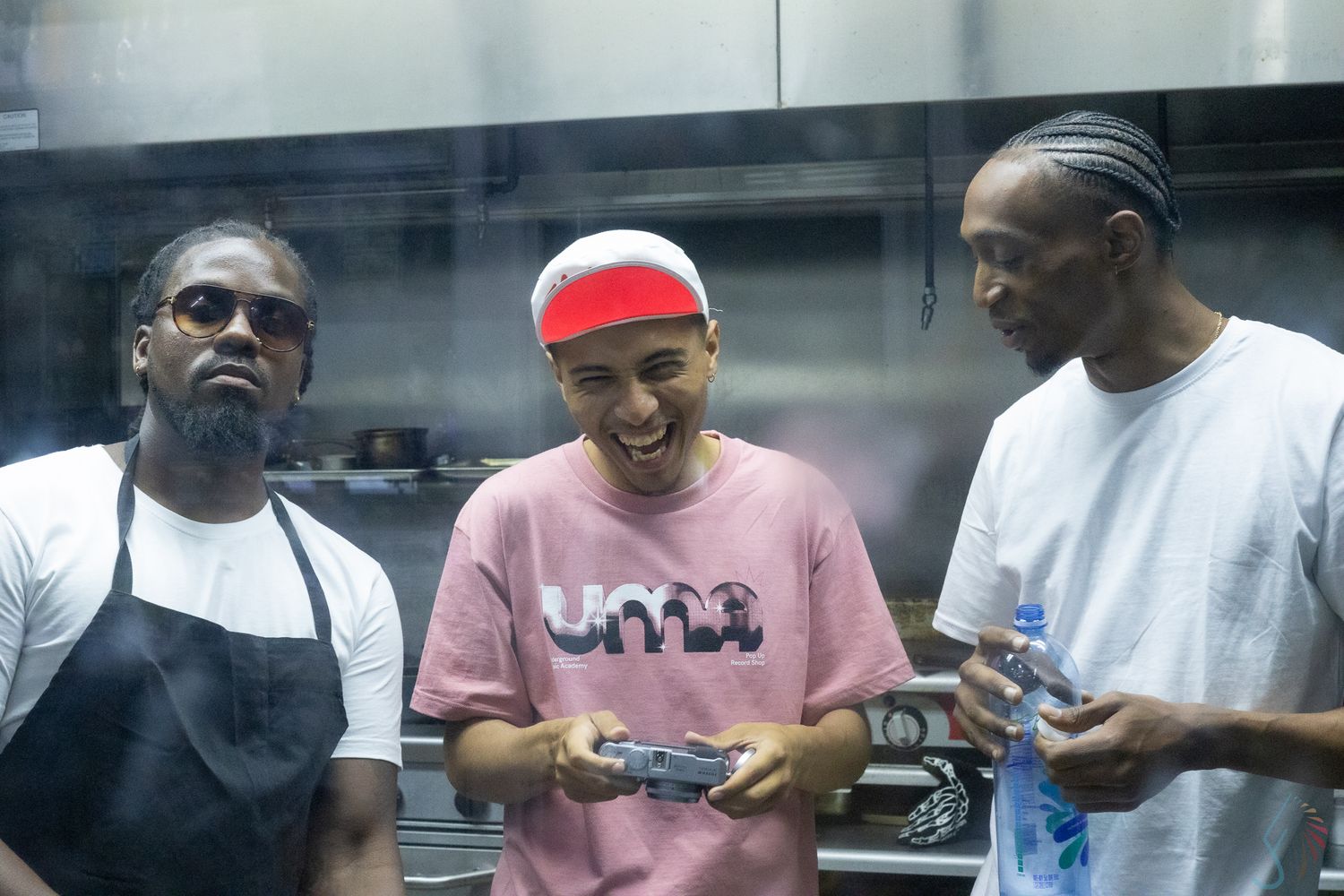 Three men smiling and laughing together in a kitchen, one wearing an apron.