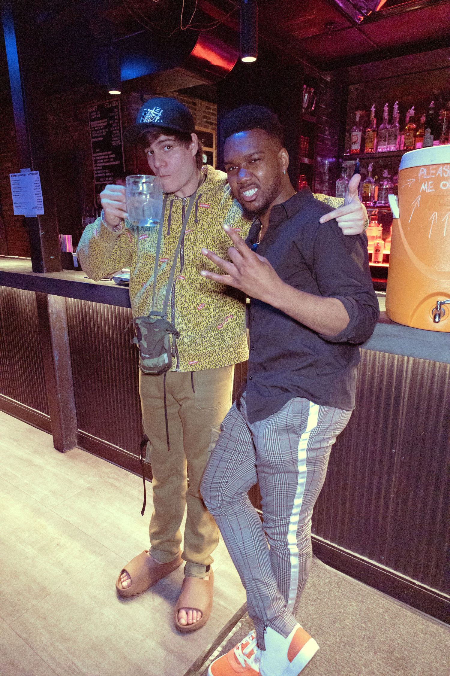 Two young men pose at a bar, one holding a large drink while the other gives a thumbs up in casual streetwear.