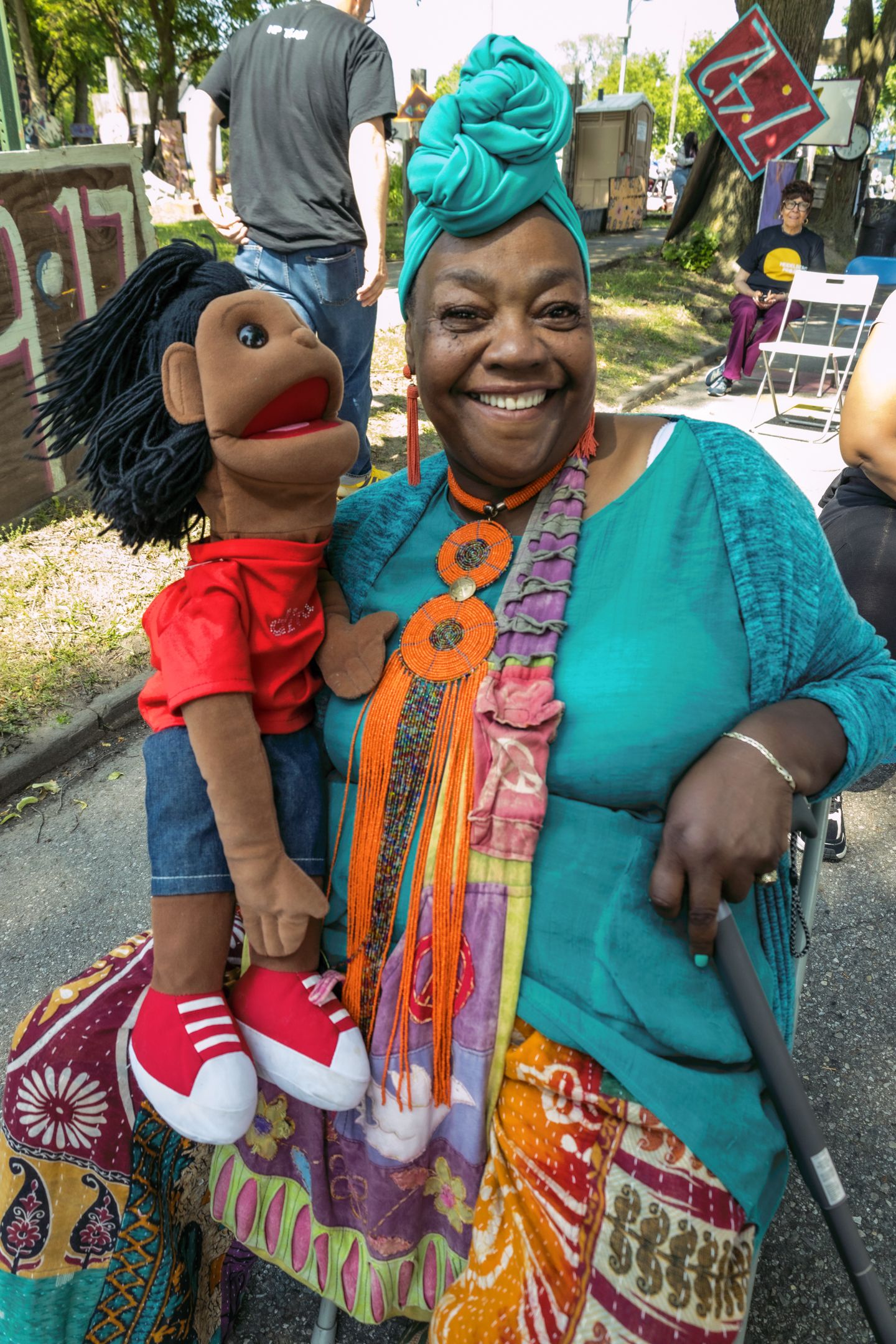 Smiling woman in teal headwrap and colorful outfit holds a puppet at an outdoor festival.