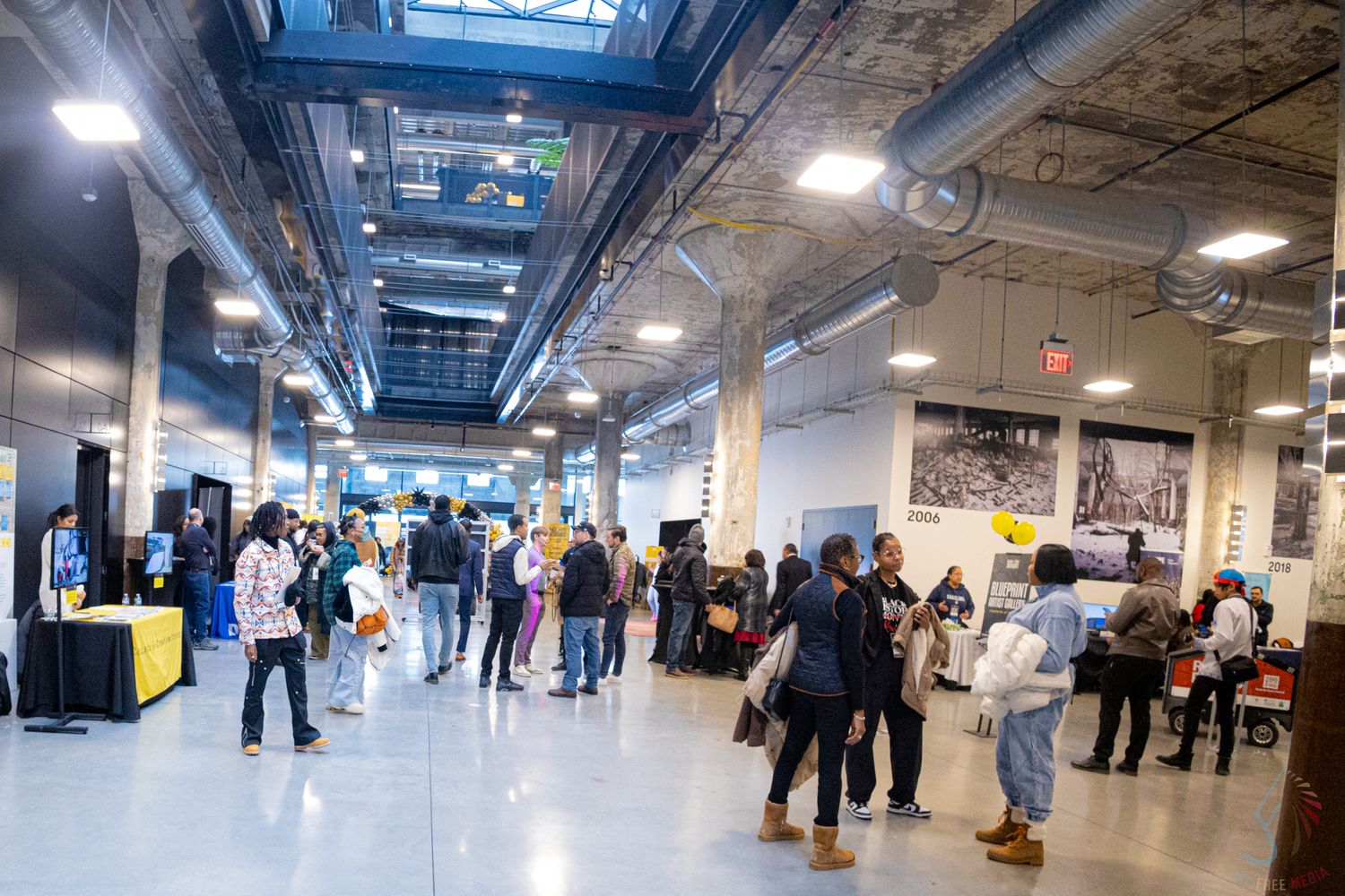 Crowd of diverse attendees browsing exhibits at an indoor trade show or art fair in a modern industrial venue.