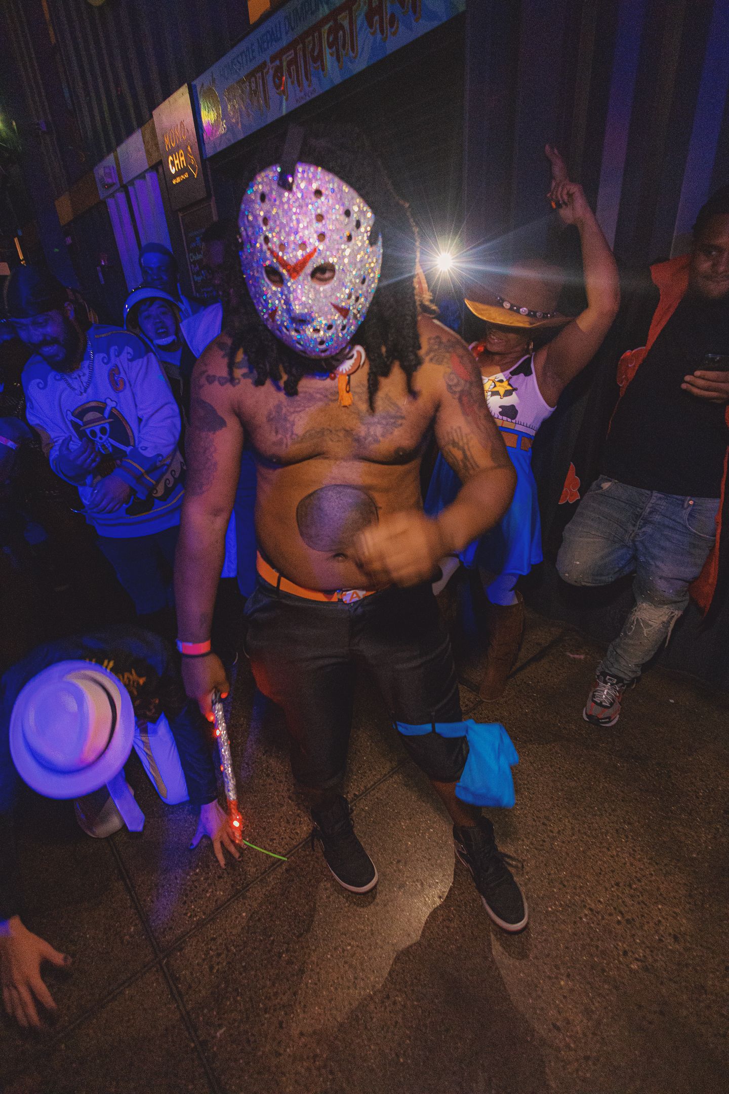 Shirtless man in hockey mask costume dancing at Halloween party under blue and purple lights.