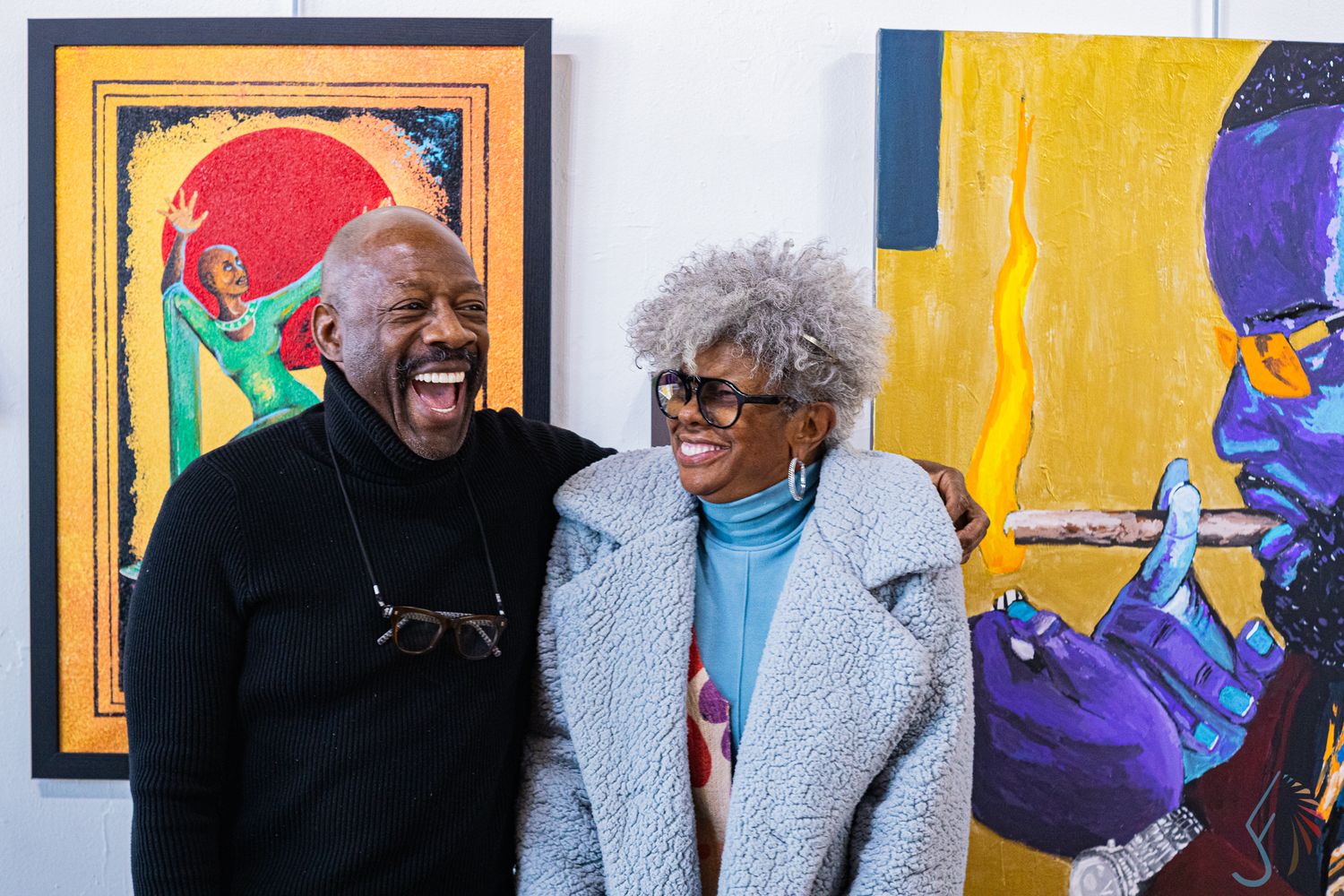Two people laughing joyfully in front of colorful African American artwork paintings at an art gallery.