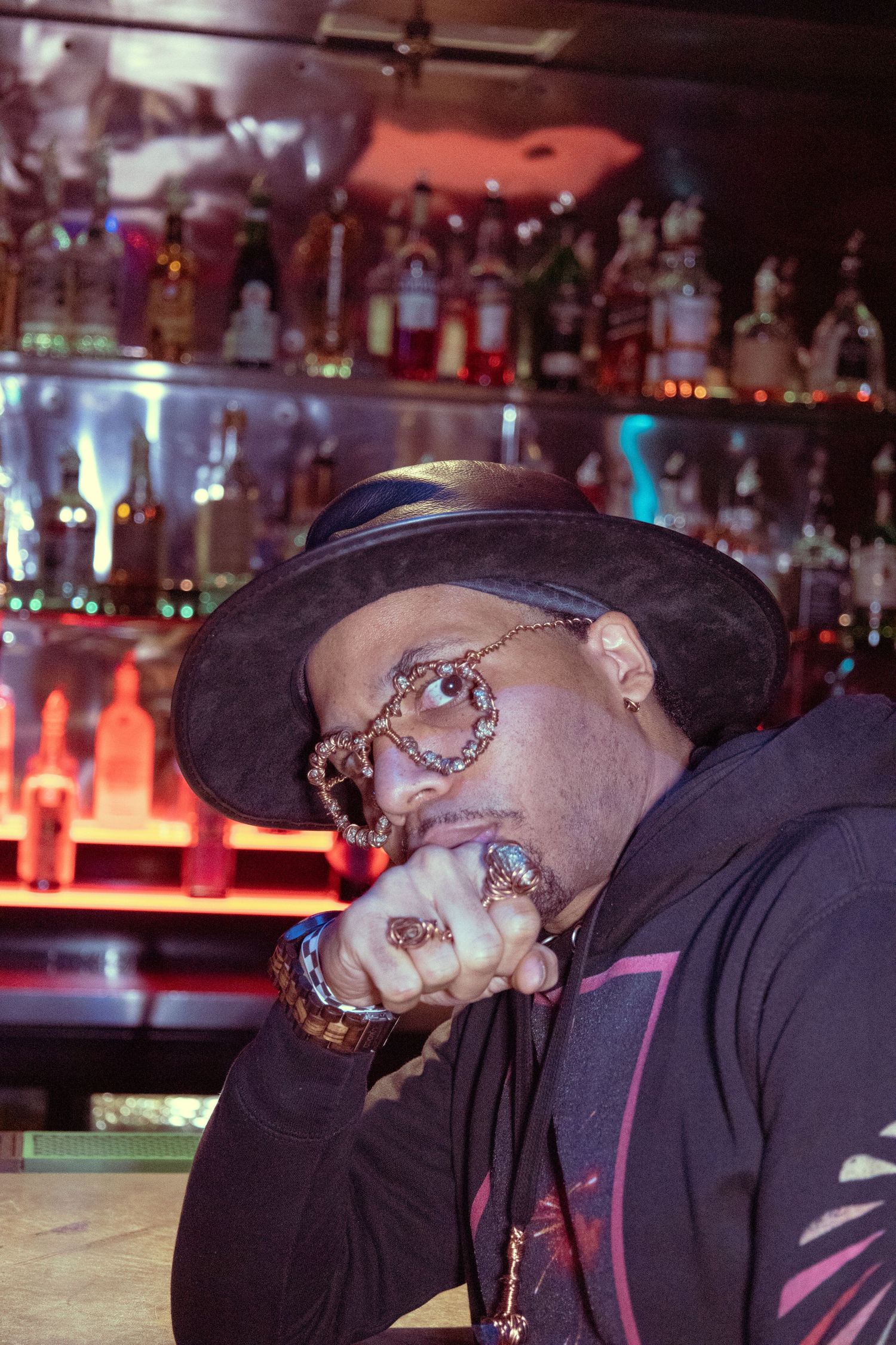 A stylish man poses at a bar wearing a black hat and ornate steampunk-style circular glasses with gold rings.