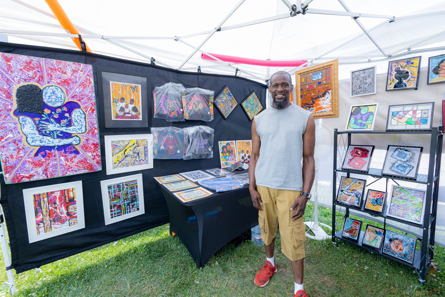 Black man in gray sleeveless shirt poses at outdoor art festival booth showcasing vibrant colorful paintings and prints.
