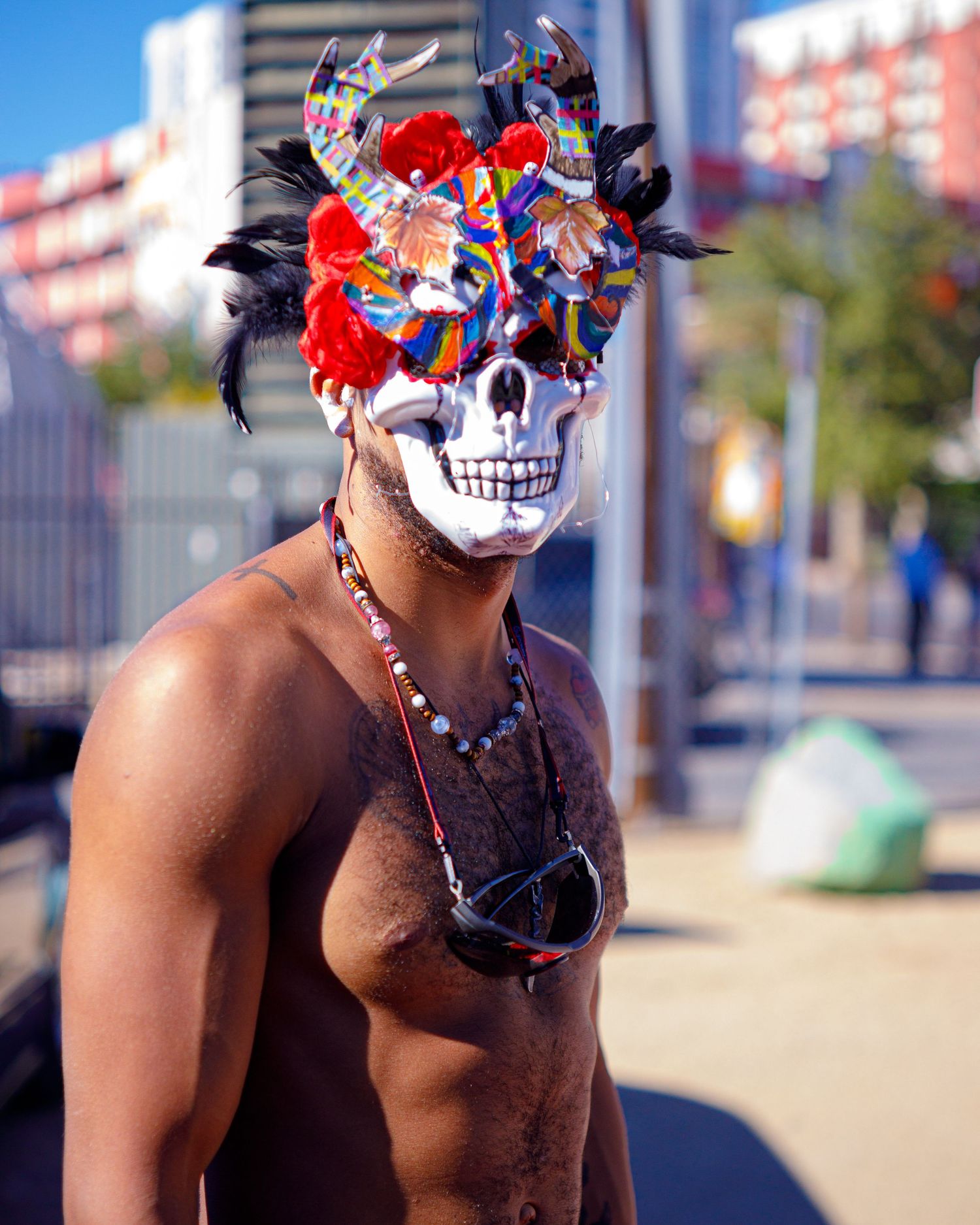 Person wearing a decorative skull mask with red feathers outdoors on a sunny day.