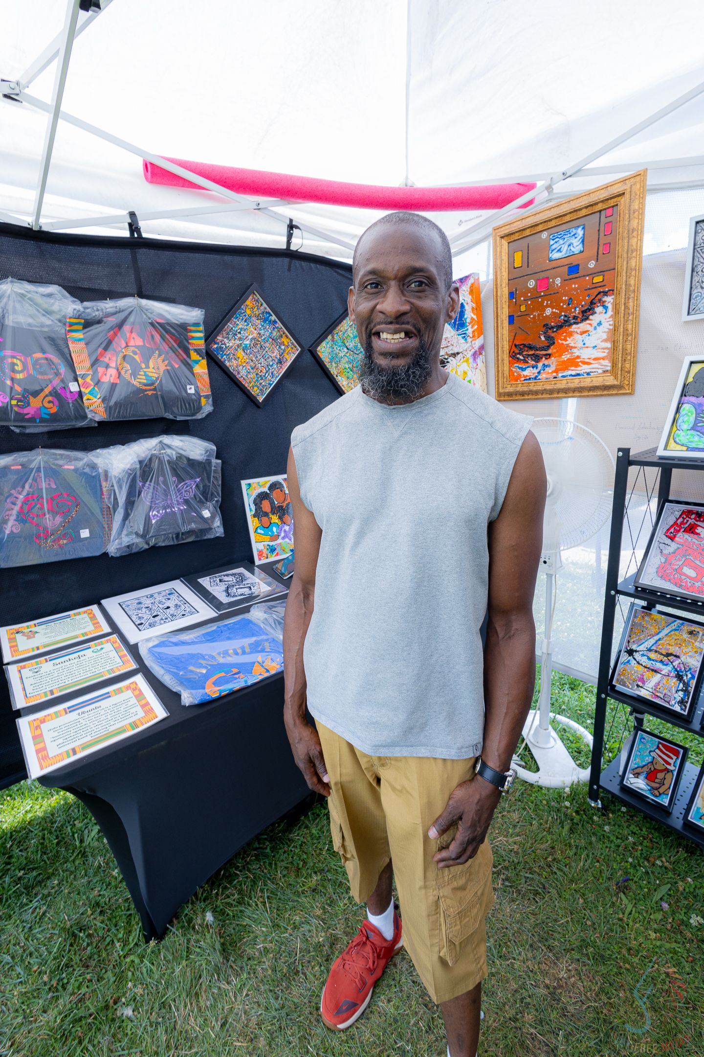 Smiling Black man in gray sleeveless shirt and khaki shorts stands at outdoor art fair booth displaying colorful artwork.