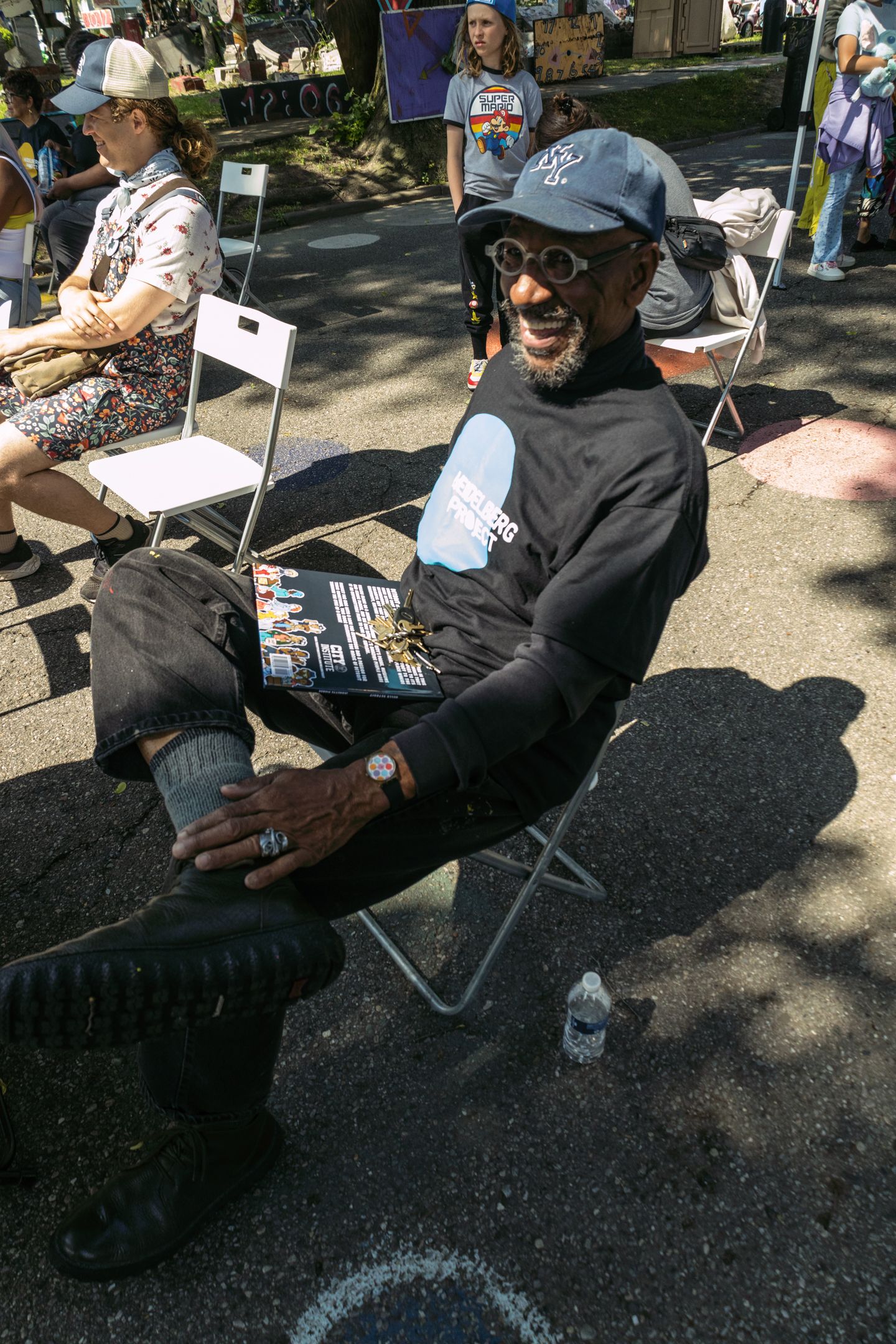Man in black hoodie and cap relaxes in folding chair outdoors at sunny community event.