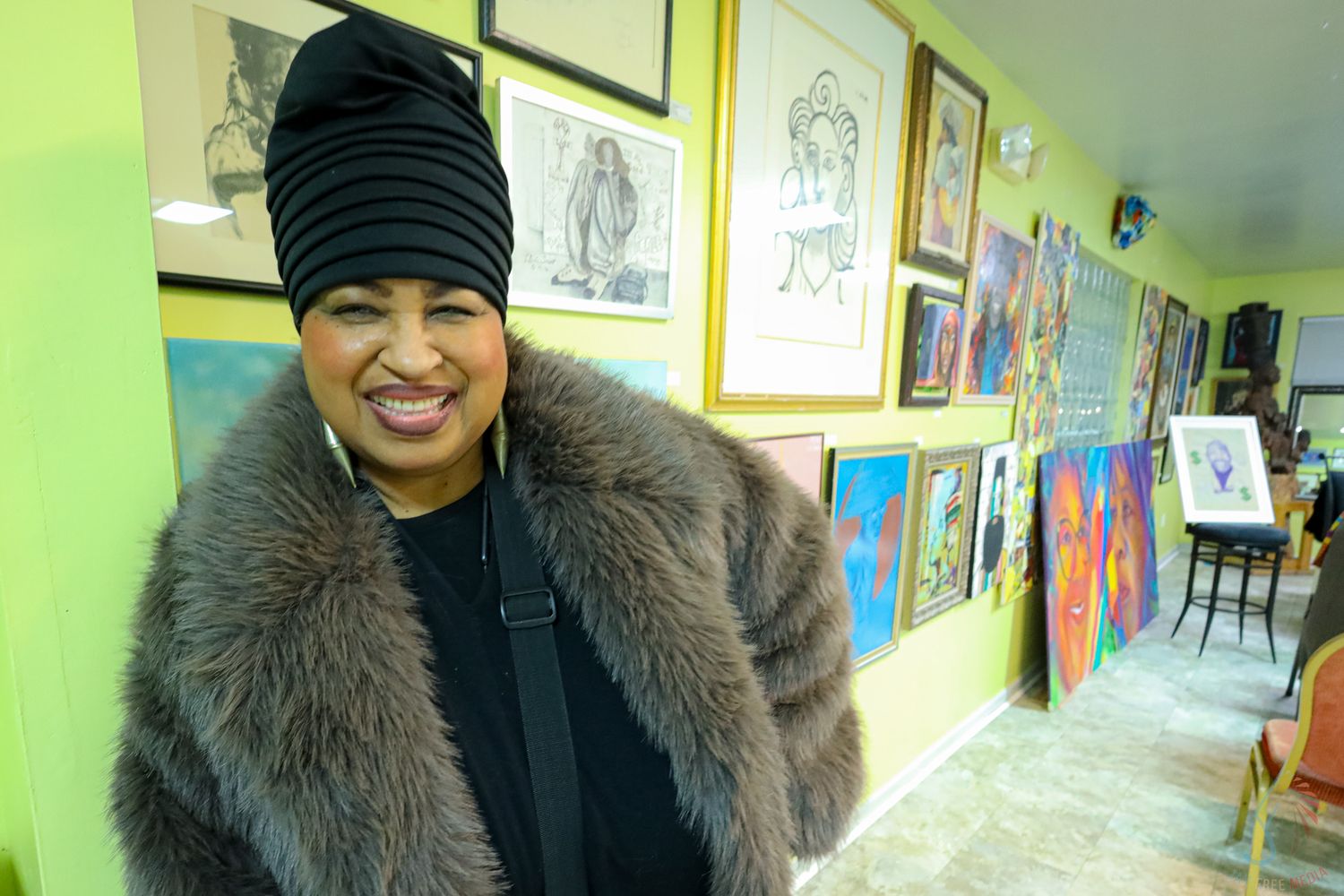 Woman in black turban and fur coat smiling in colorful art gallery with paintings on yellow walls.