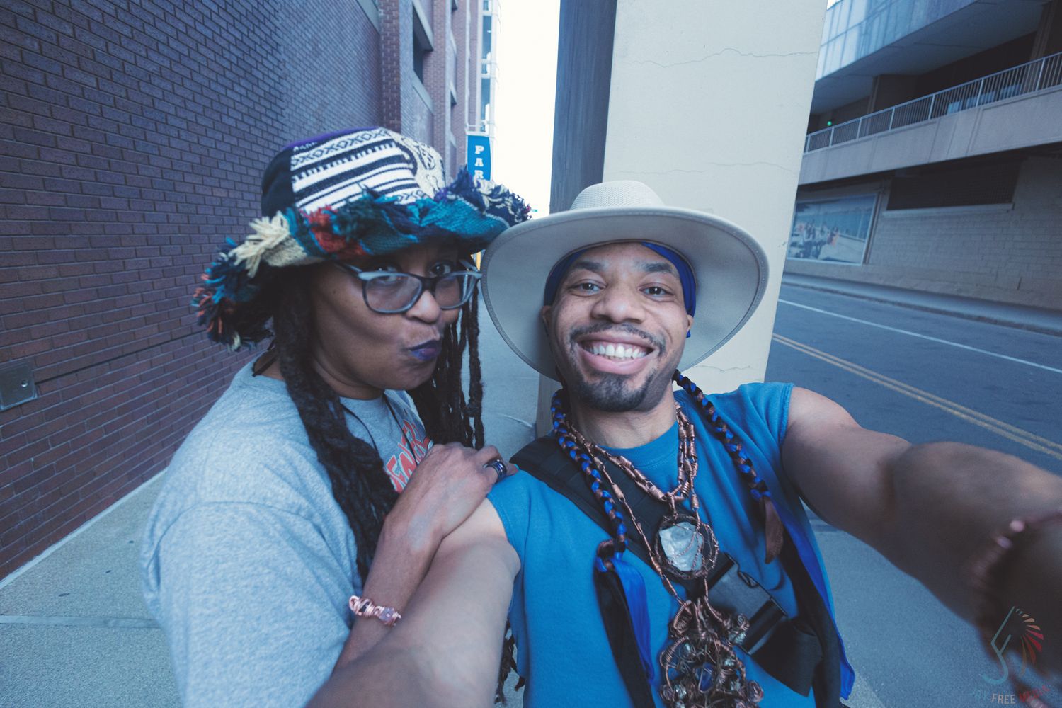 Two smiling friends taking a selfie outdoors in an urban alley, wearing stylish hats and blue outfits.