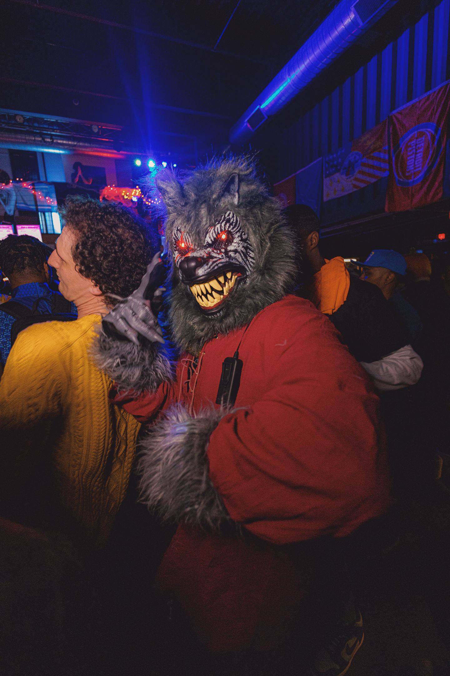 Person in scary werewolf Halloween costume with glowing red eyes at a crowded party event.