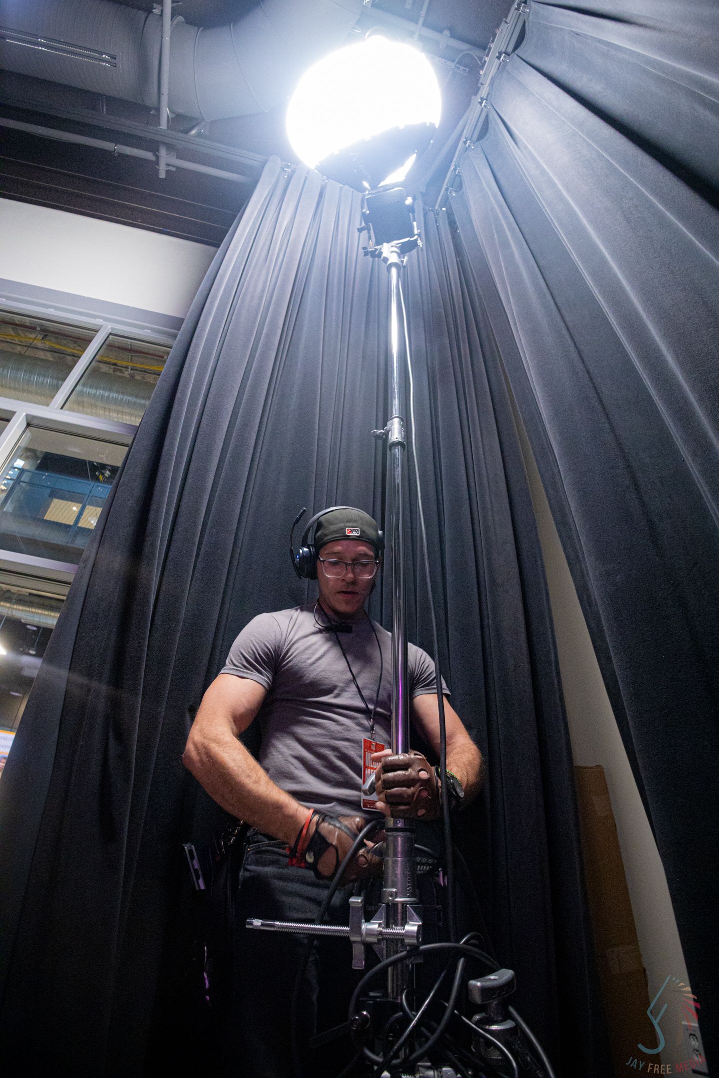 A muscular man in a gray shirt operates a tall light stand with a large globe light in a studio setting.