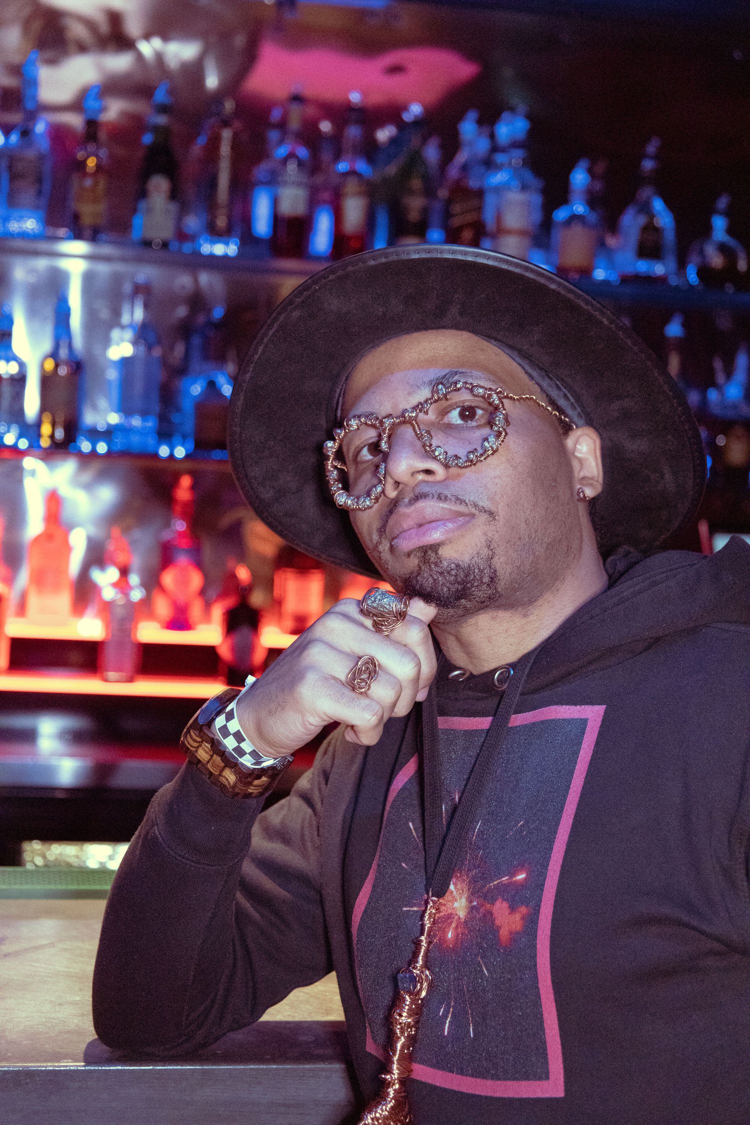 A stylish man wearing a wide-brim hat and decorative glasses poses at a bar with colorful bottle-lined shelves behind him.