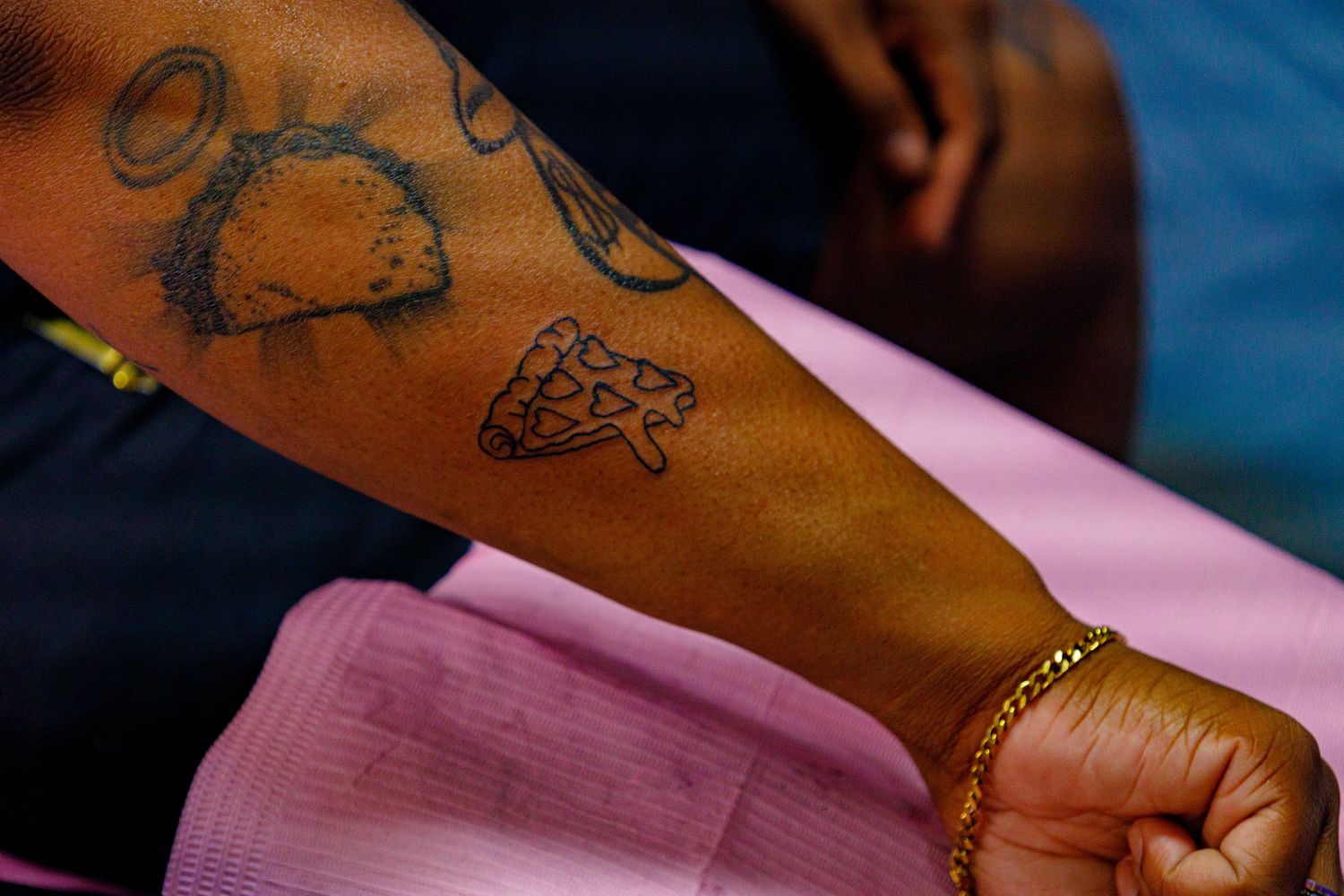 Close-up of tattoos on a dark-skinned arm, featuring a pizza slice and taco designs, with a gold bracelet.