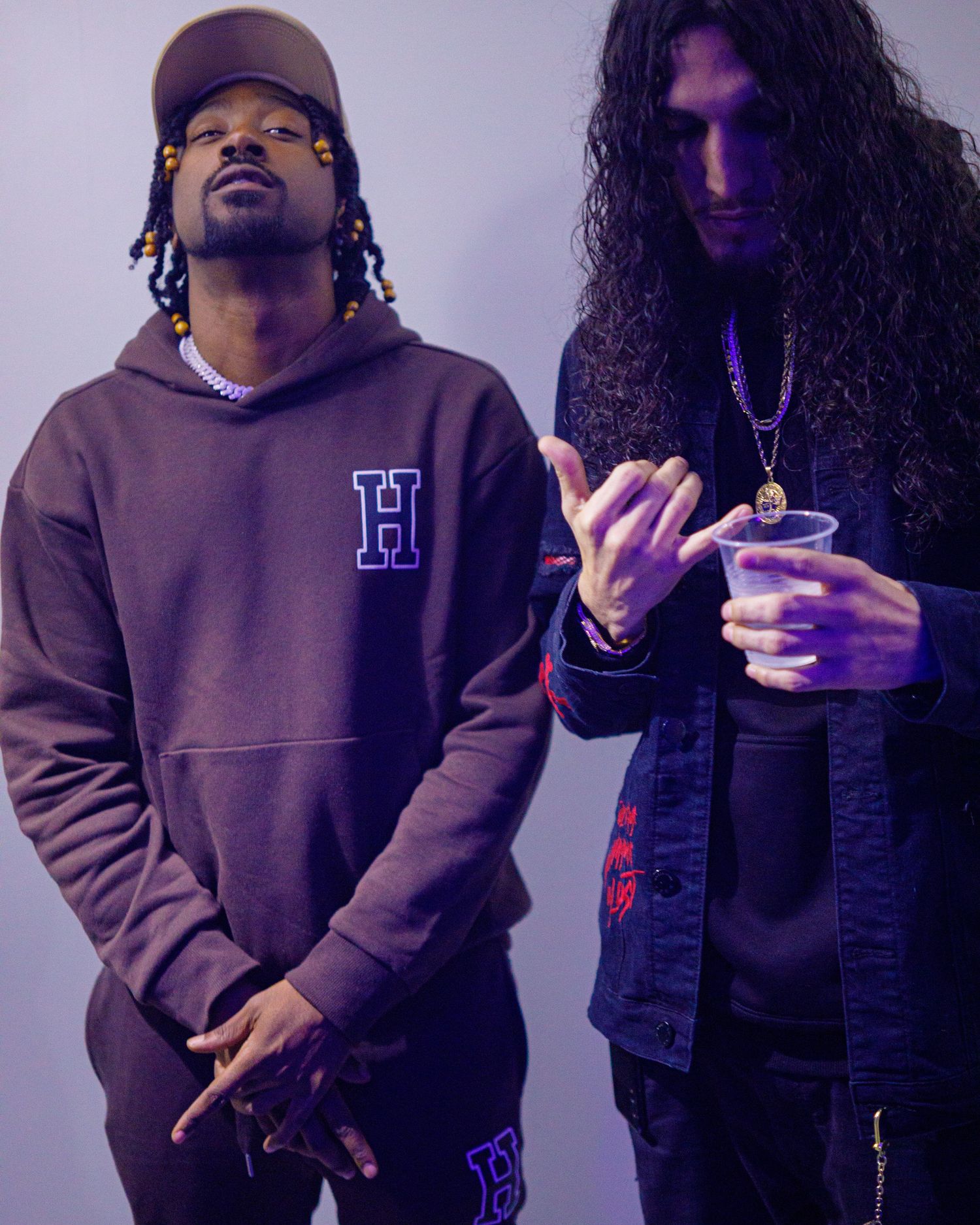 Two men in dark streetwear posing together under purple studio lighting.