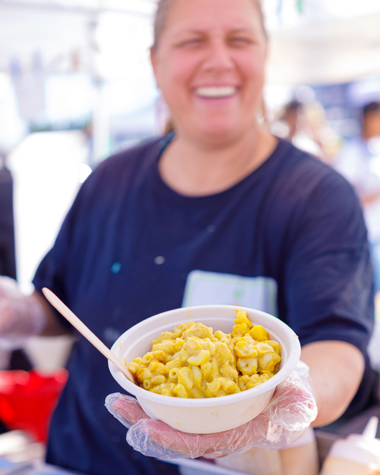 Smiling vendor holding bowl of yellow mac and cheese at outdoor food festival booth.