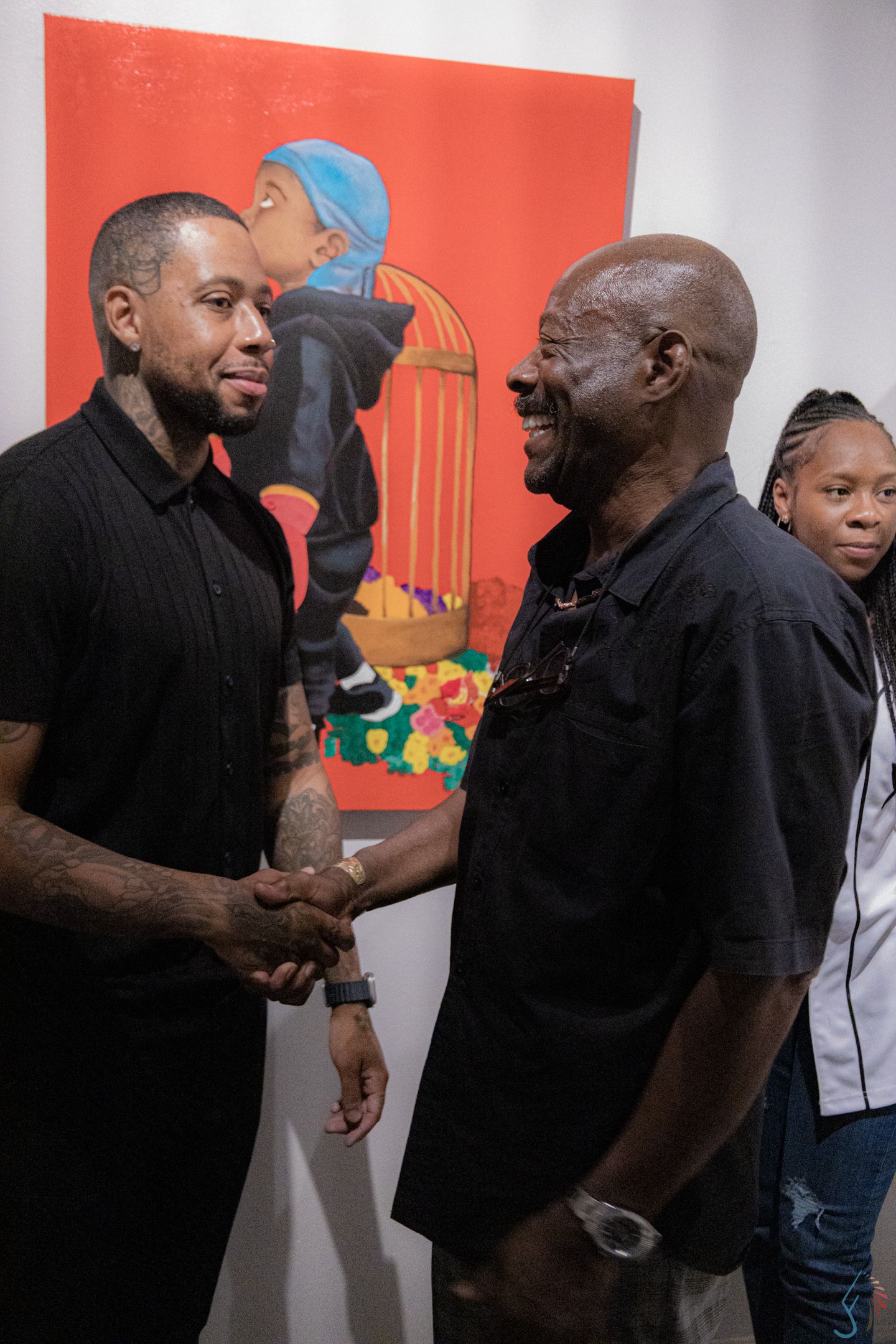 Two men shake hands warmly at an art gallery with a bold red painting displayed on the wall behind them.