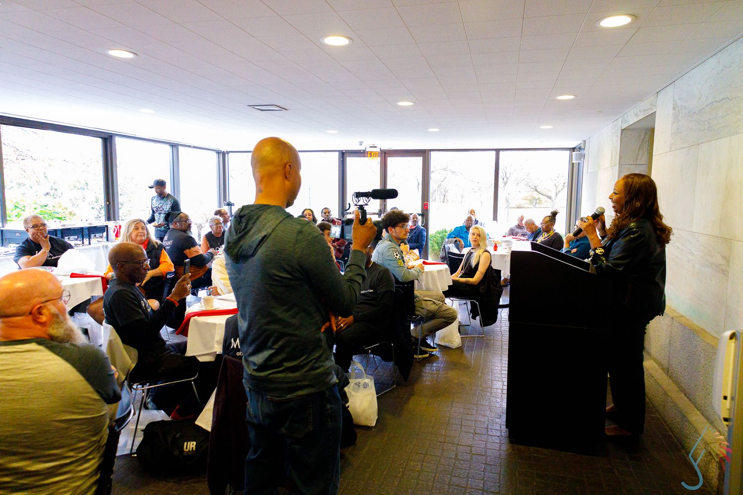 Audience seated at indoor event listening to speaker at podium in modern conference room with large windows.