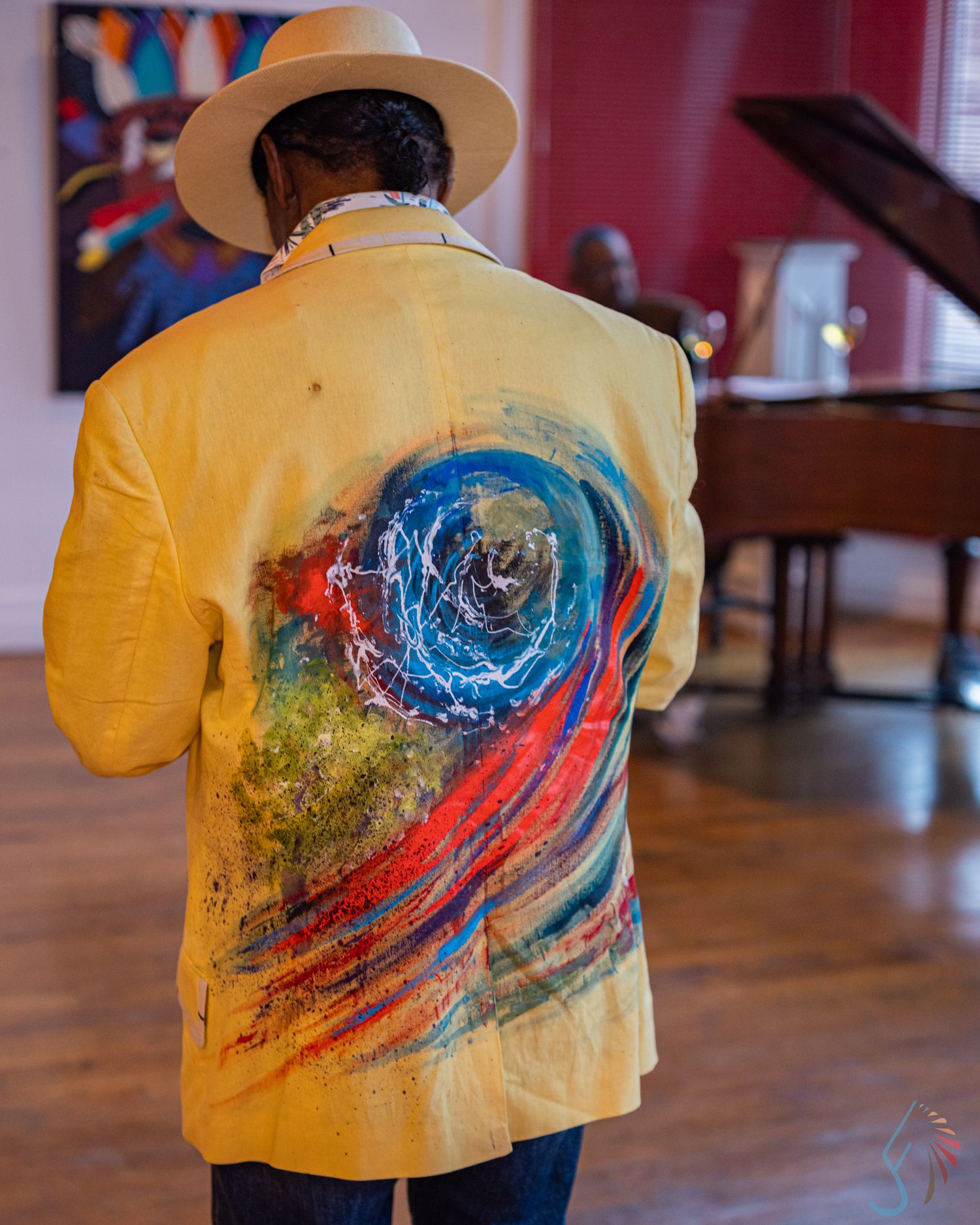 Man in white hat and yellow painted blazer featuring colorful swirl art stands near a grand piano.