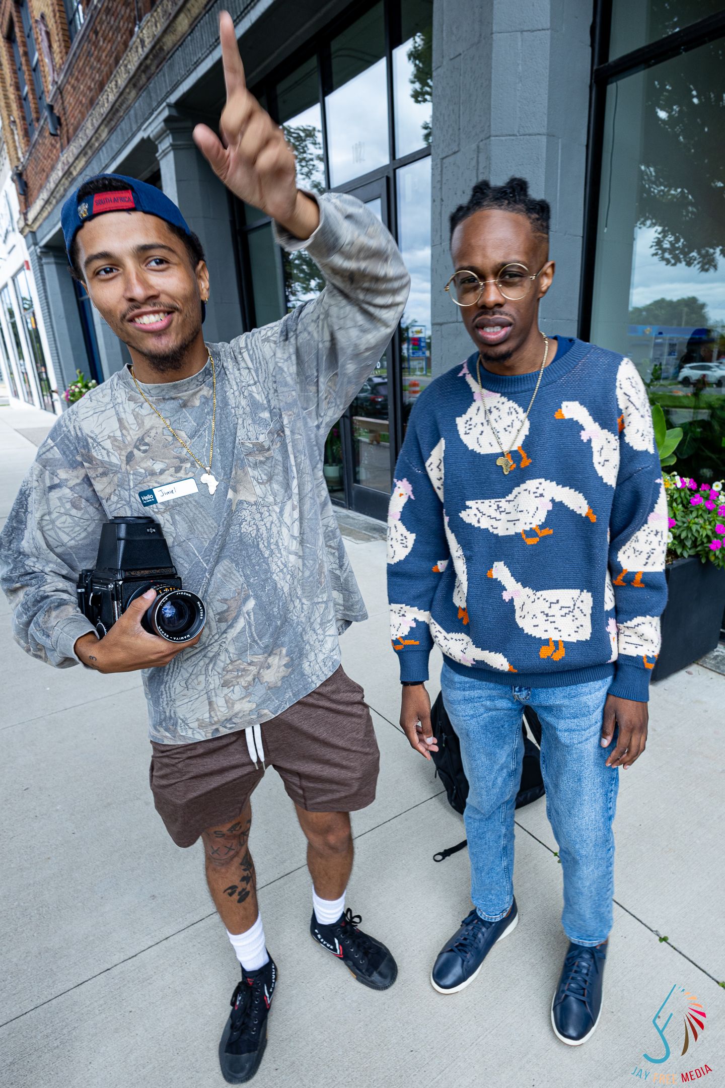 Two stylish young men posing on a city sidewalk, one holding a camera and pointing upward.