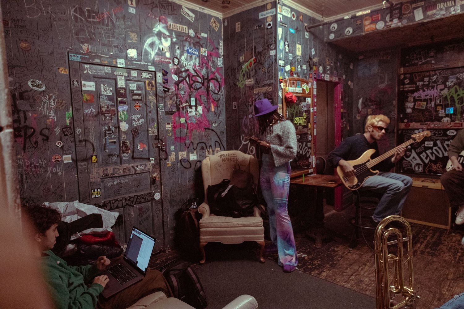 Musicians jamming in a graffiti-covered room with colorful stickers, one playing guitar while another uses a laptop.
