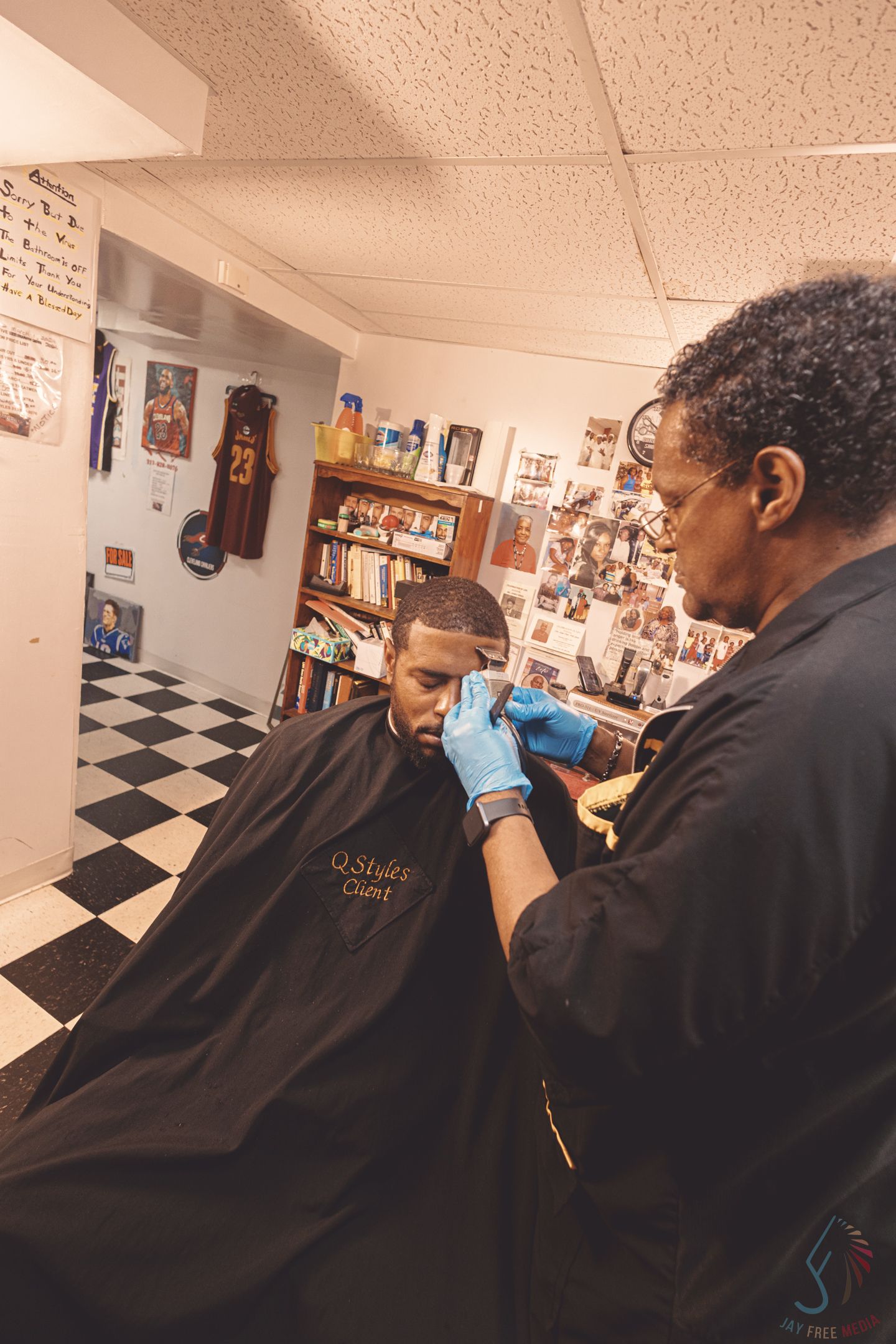 Barber cutting a young man's hair in a barbershop, client wearing a black cape, checkered floor visible.