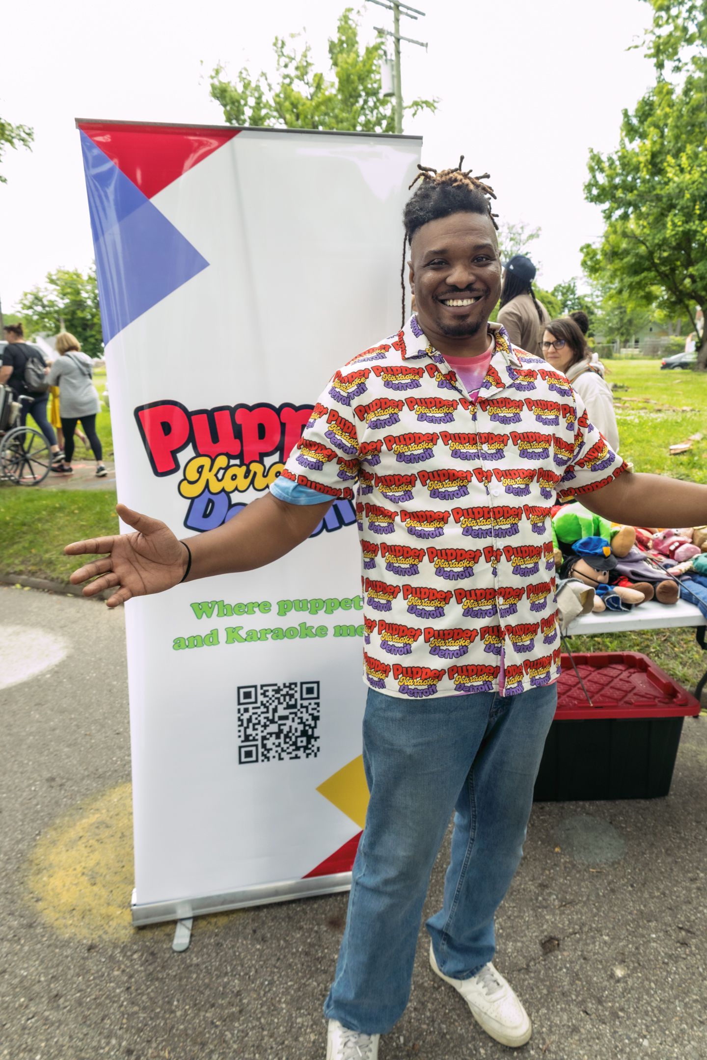 Man smiling beside a Puppet Karaoke banner at an outdoor community event, wearing a colorful patterned shirt.