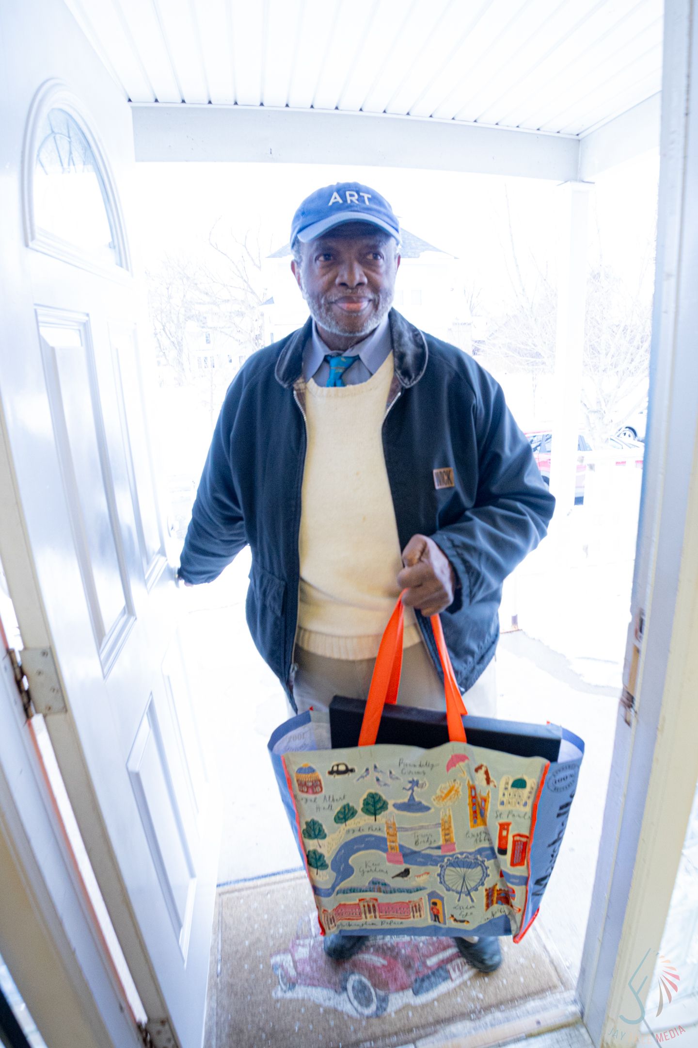 Elderly man smiling at doorway holding colorful reusable shopping bag, wearing blue cap and jacket.