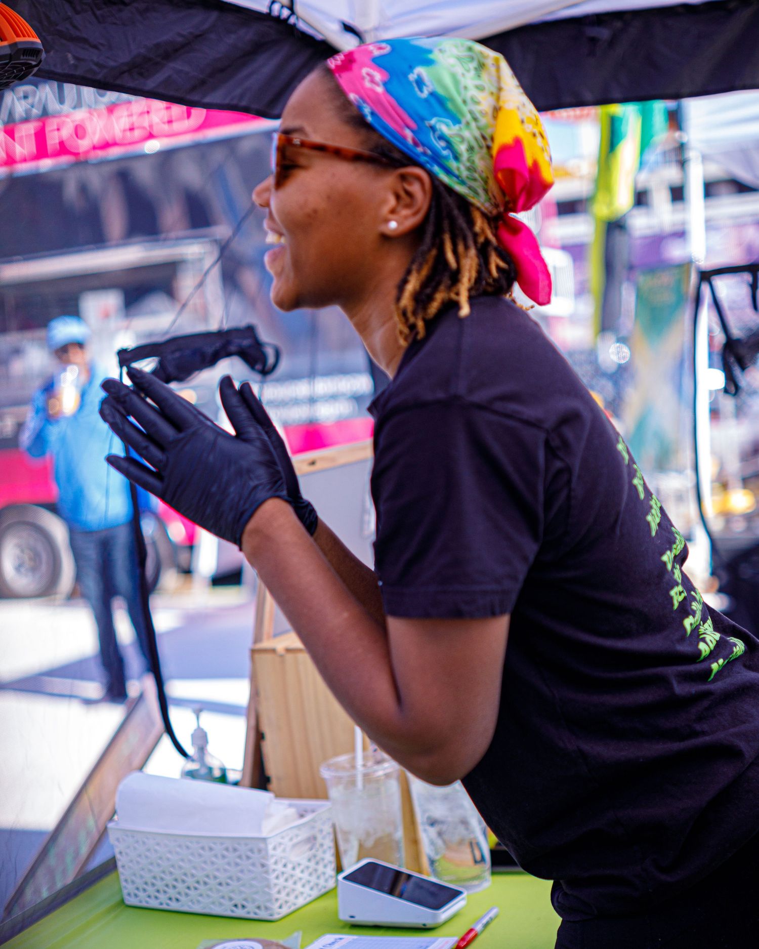 Woman in colorful headscarf and black gloves working at an outdoor food vendor booth.
