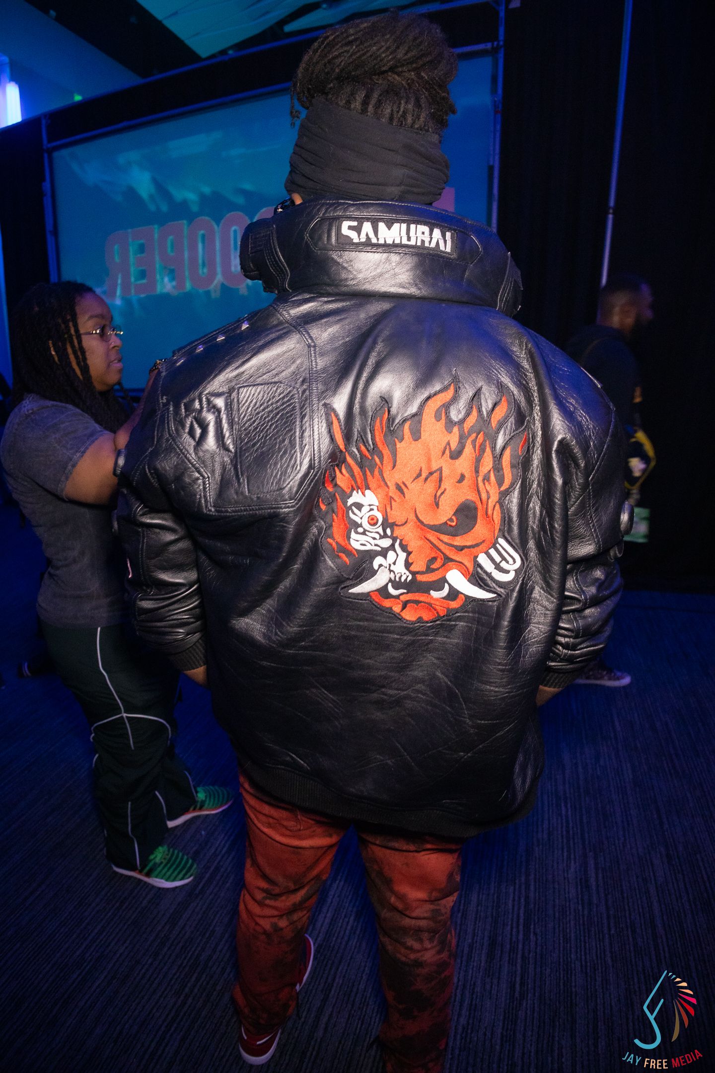 Person wearing a black leather jacket with a flaming skull graphic and Samurai hat at a neon-lit event.