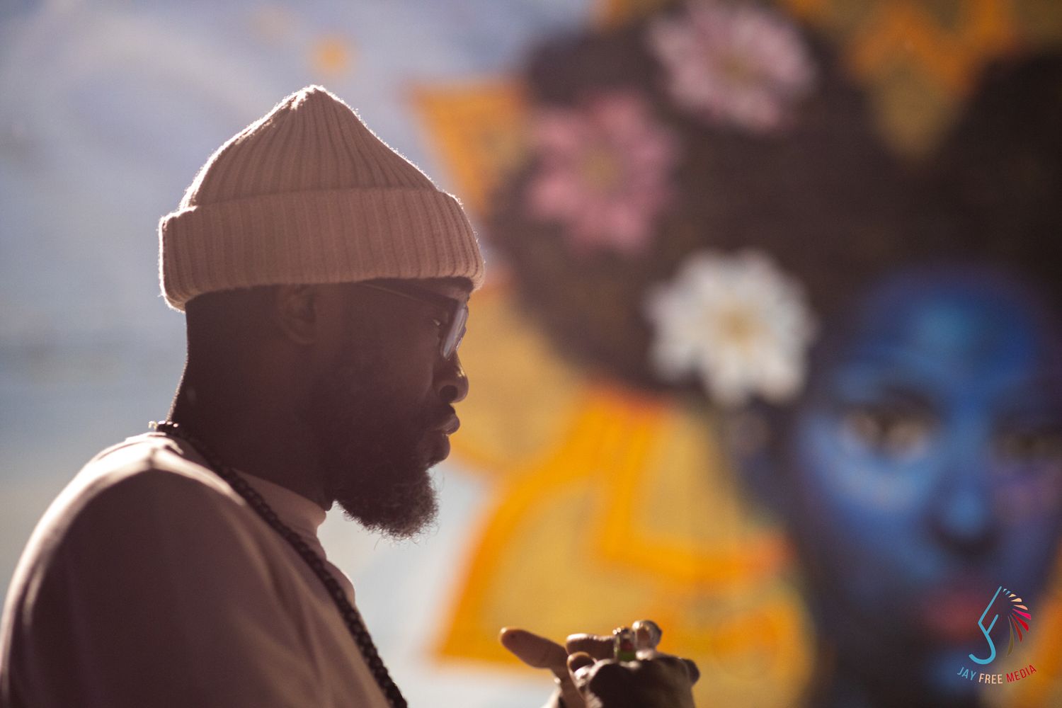A bearded Black man in a beige beanie stands before a colorful mural featuring a blue-faced woman with flowers.