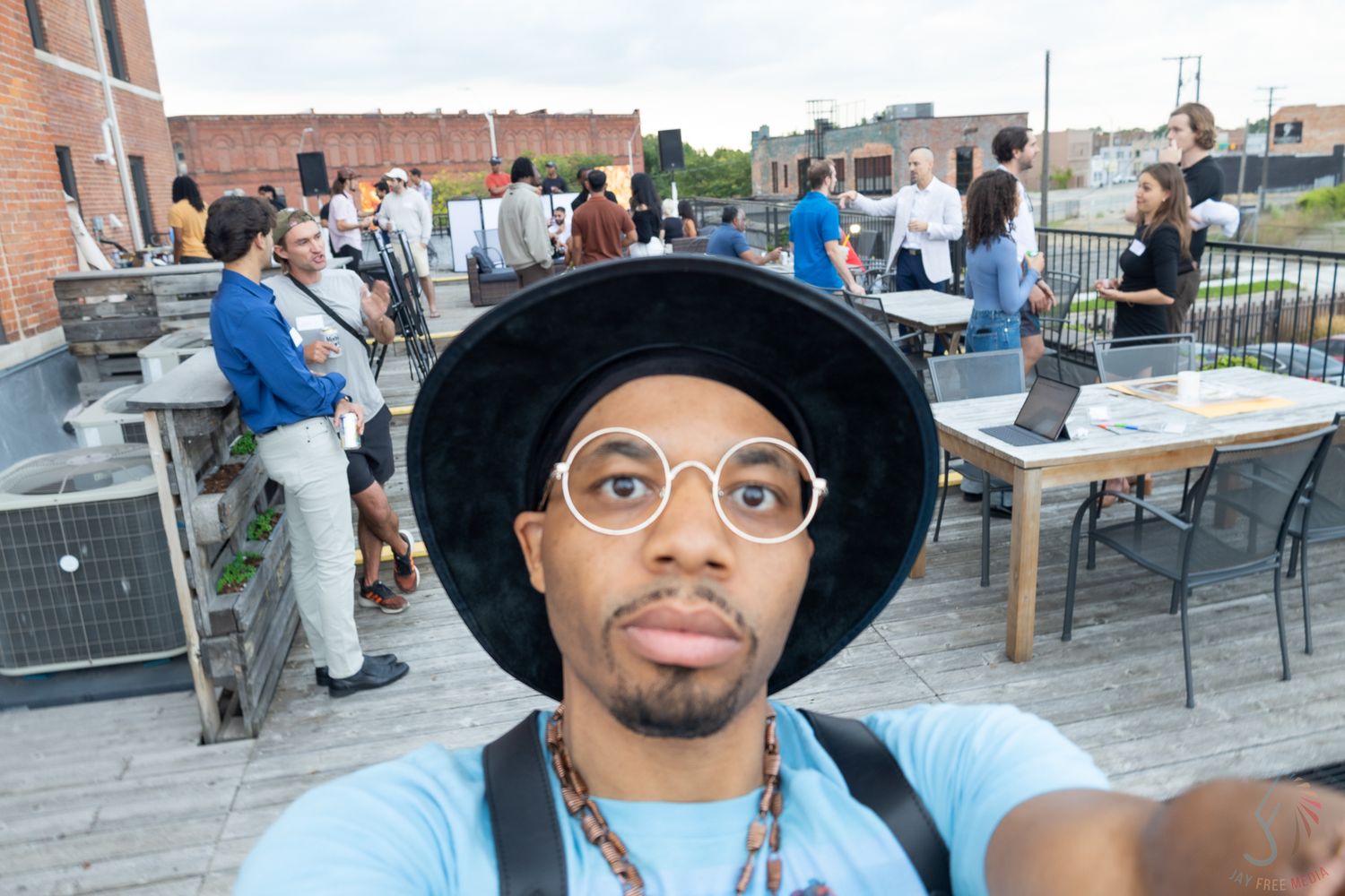 Man in black hat and glasses takes rooftop selfie at outdoor social gathering event.