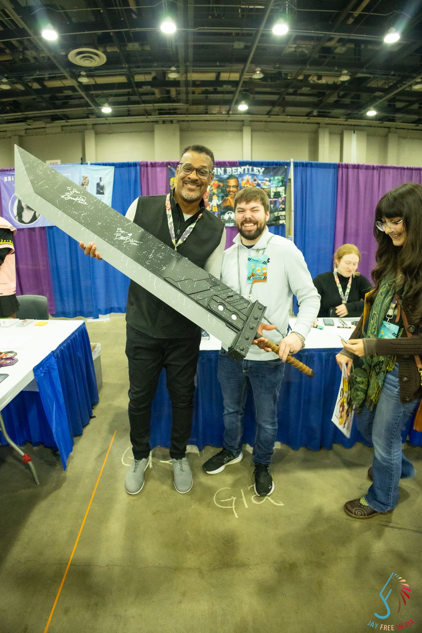 Two men pose with large signed fantasy sword prop at comic convention booth with purple backdrop.