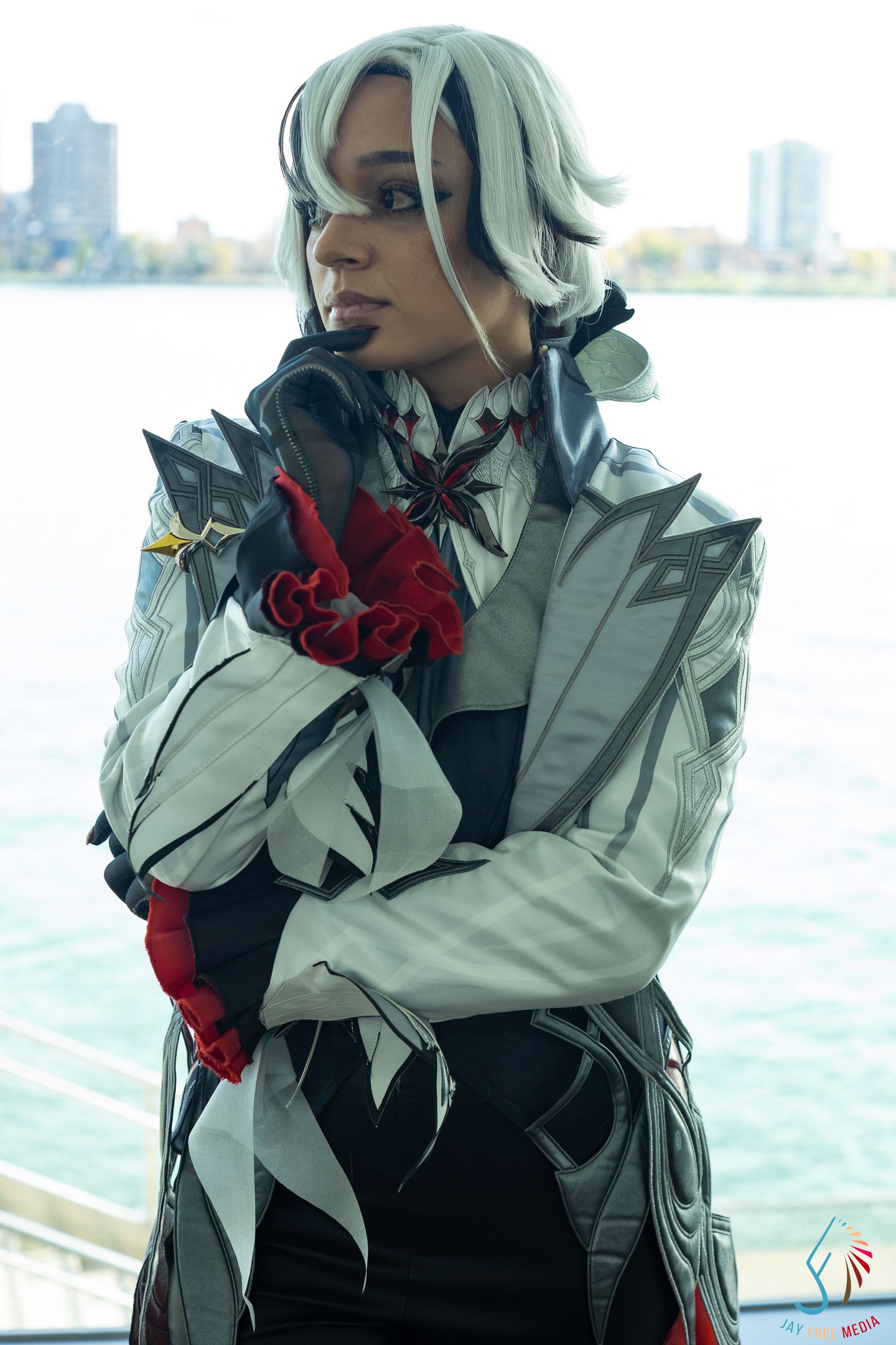 Cosplayer in white and black armored costume with red roses and silver hair at waterfront.