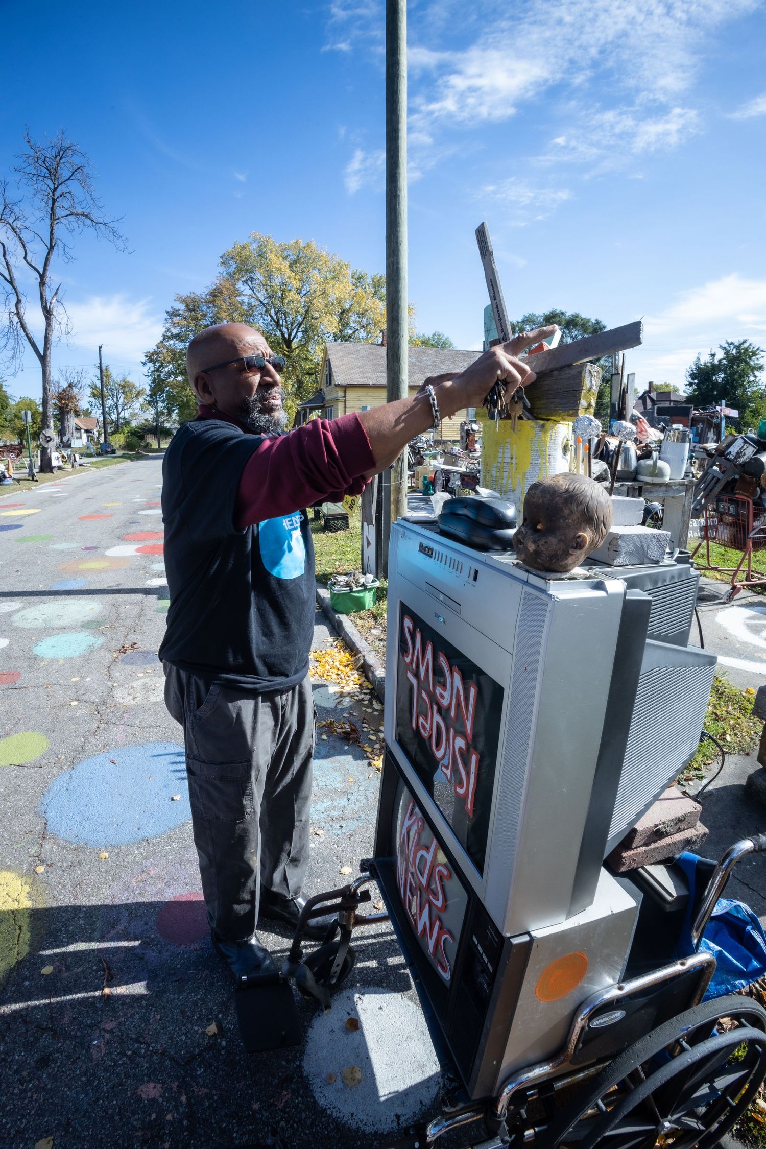 A man reaches up to arrange items on a cluttered outdoor art installation on a sunny Detroit street.