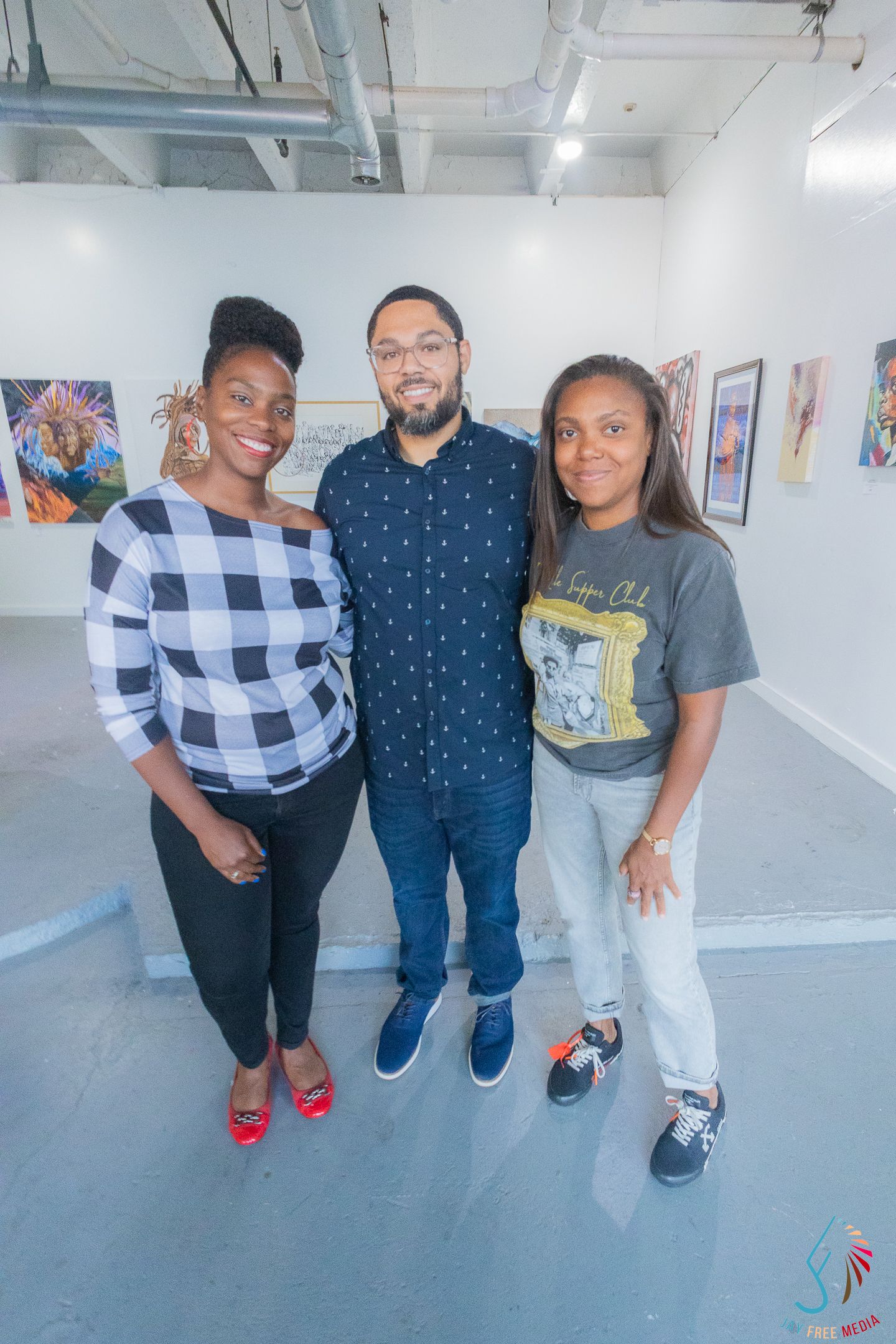 Three friends smiling together at an art gallery, surrounded by colorful artwork on white walls.