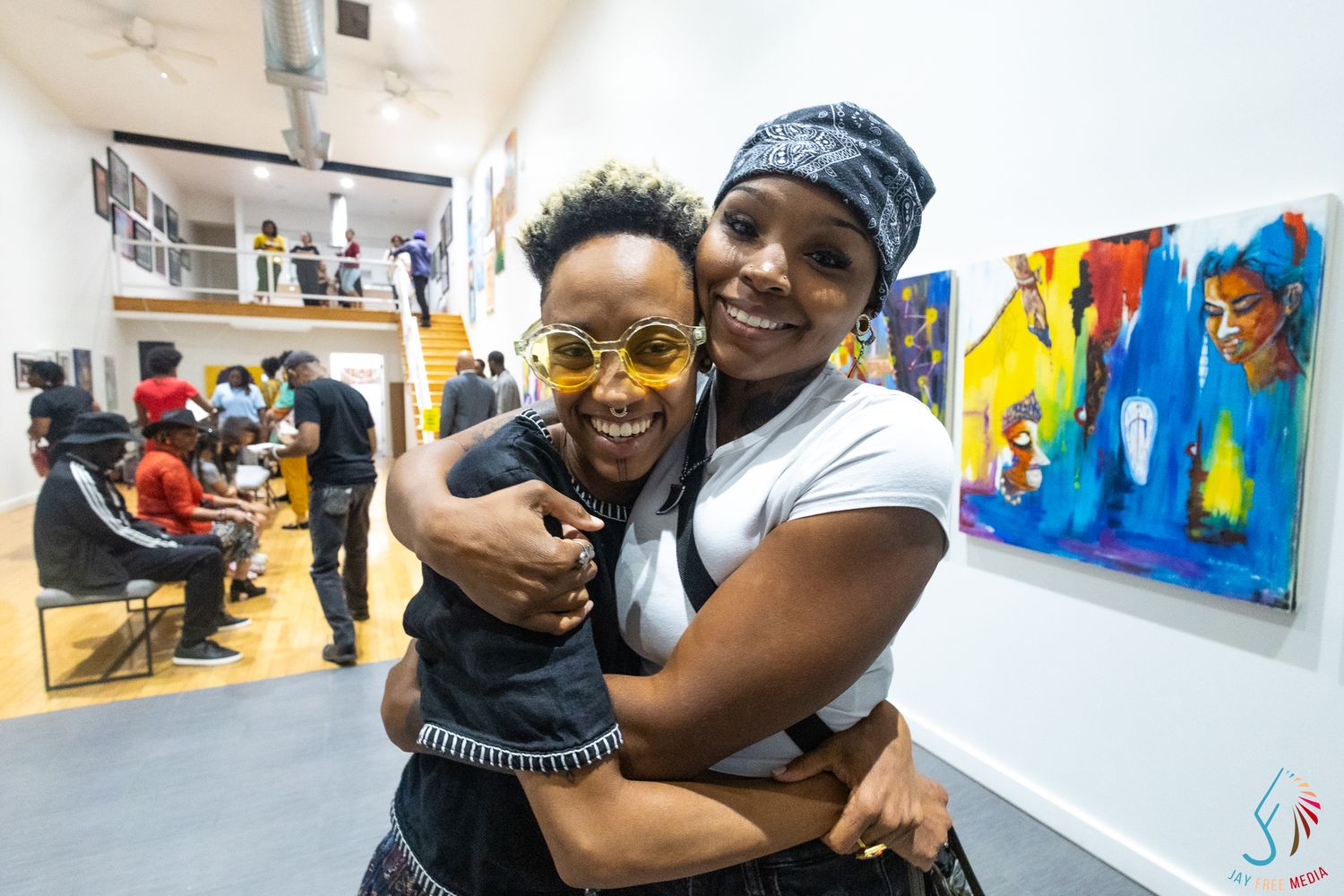 Two women share a warm hug at an art gallery event, colorful paintings displayed on white walls behind them.