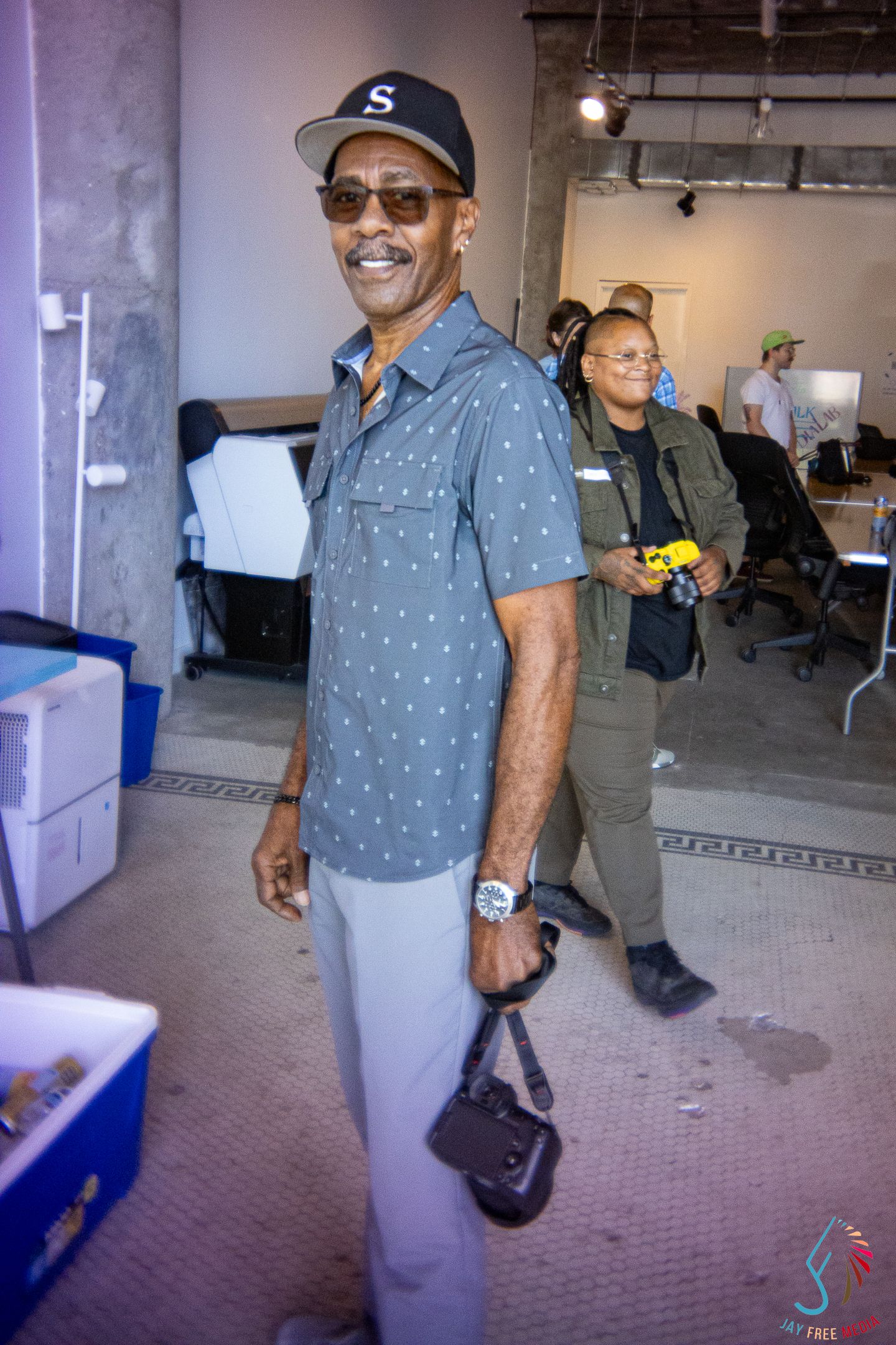 Smiling man in gray polo shirt and S cap poses indoors with a camera bag at an event.