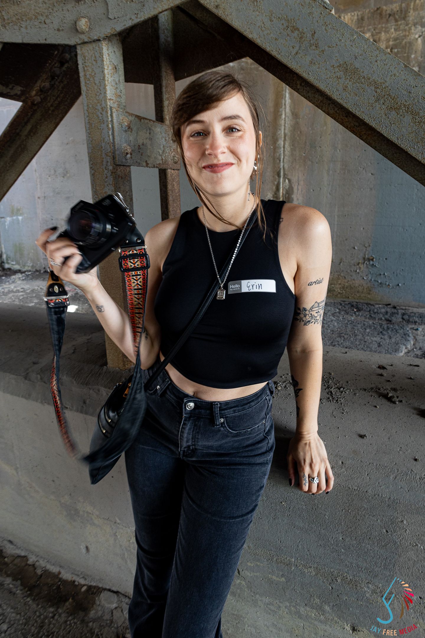 Young woman in black crop top holding a camera with a colorful strap, wearing a name tag and jeans.