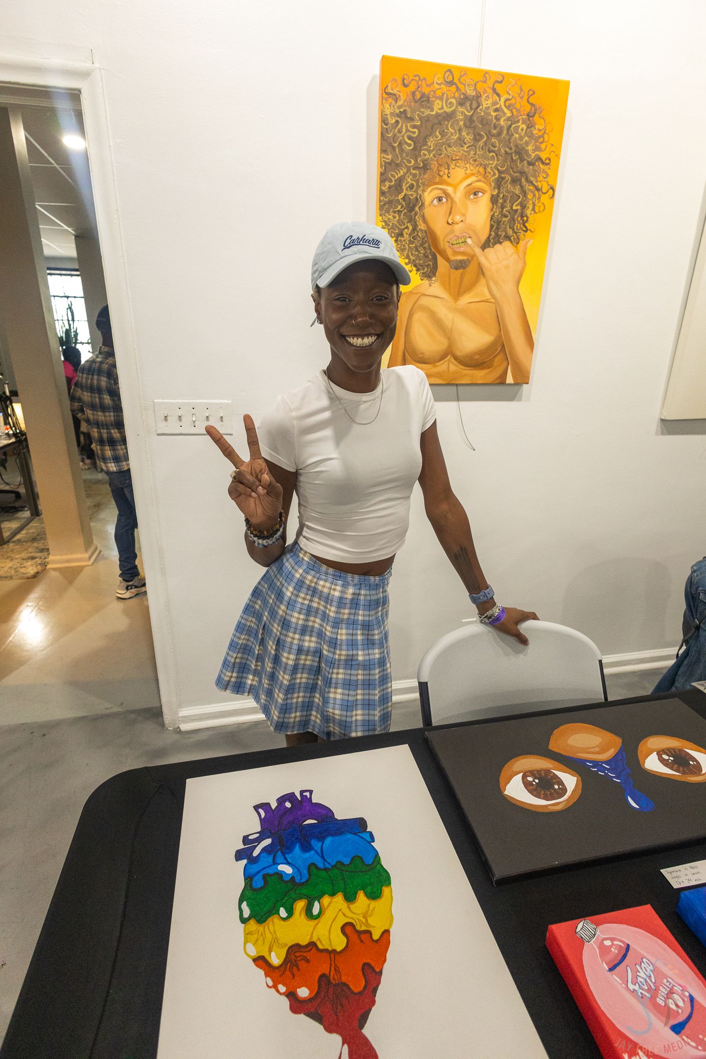 Young woman in white crop top and plaid skirt poses with peace sign at colorful art display event.