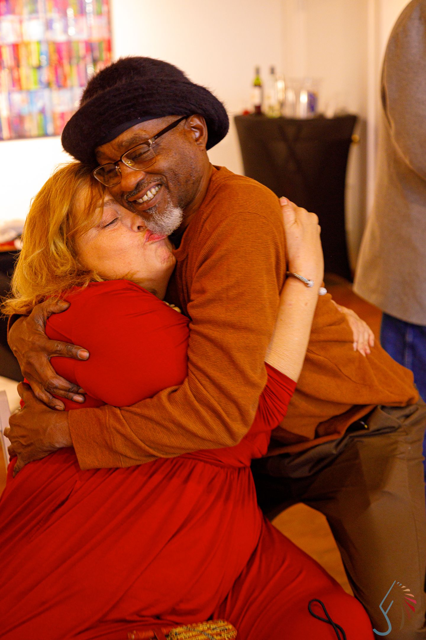 A man in a beret and woman in red share a warm embrace, smiling joyfully in an indoor setting.