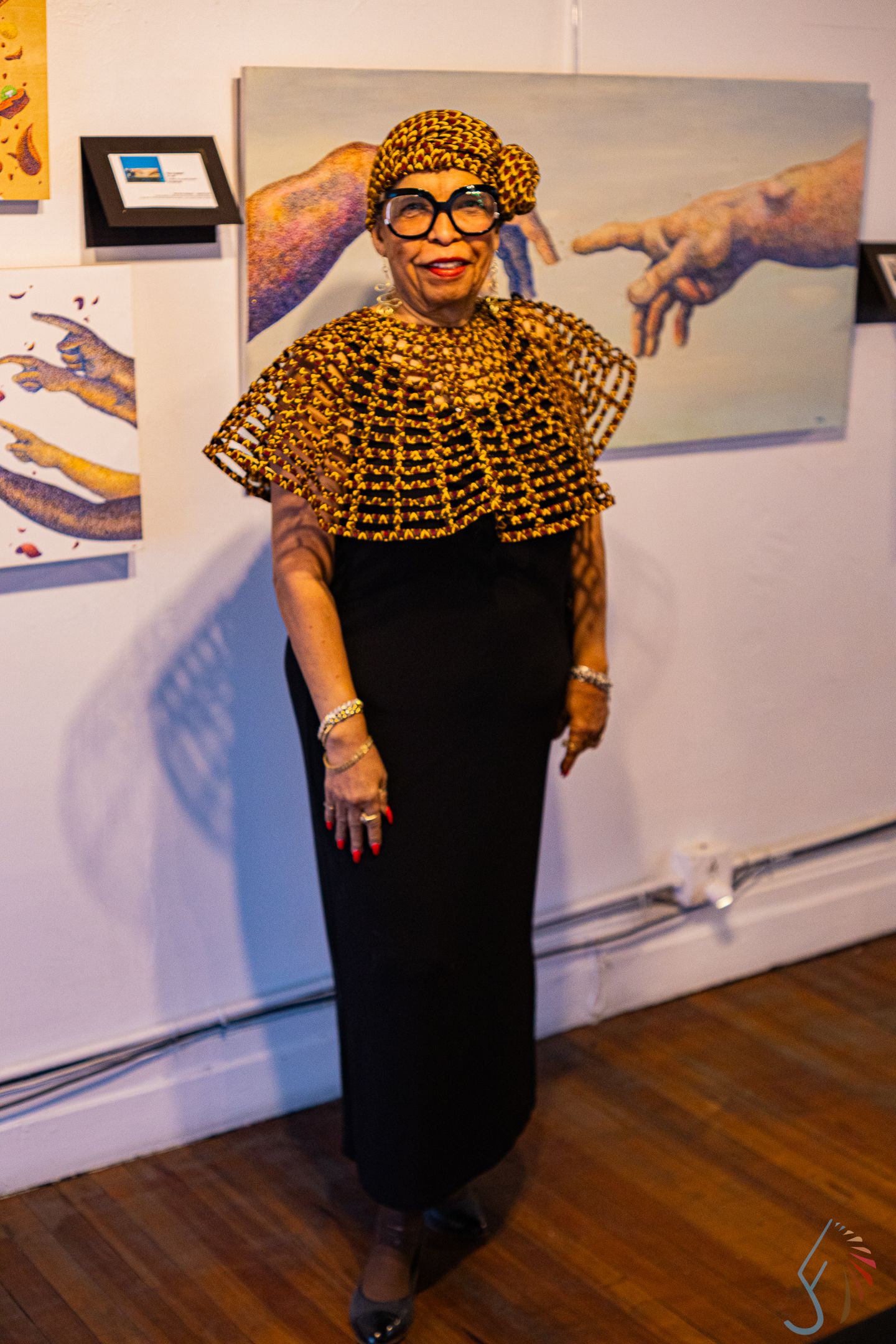 Elegant woman in black dress with gold beaded cape and matching hat poses at an art gallery exhibition.