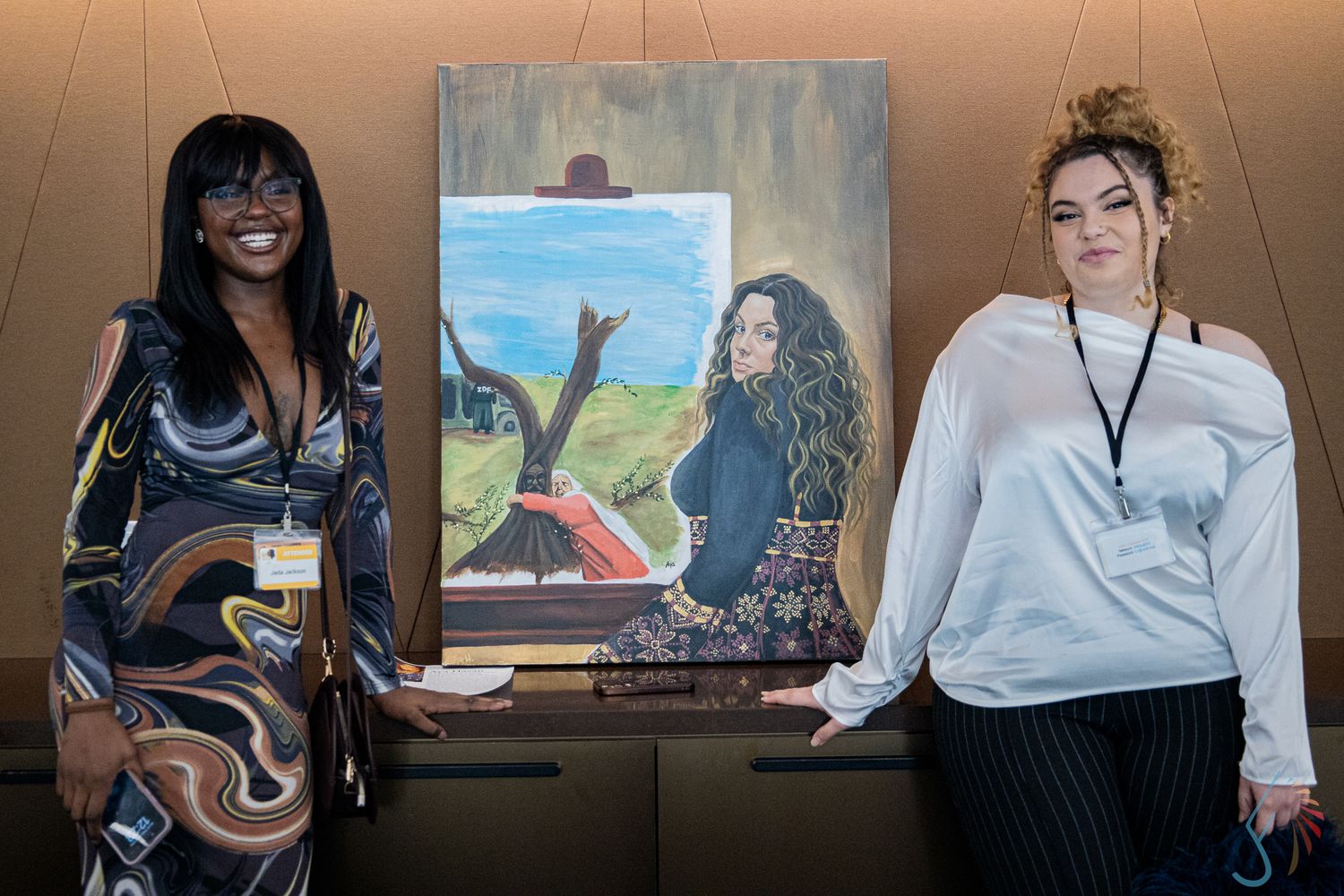 Two women standing beside a colorful portrait painting at an indoor event.
