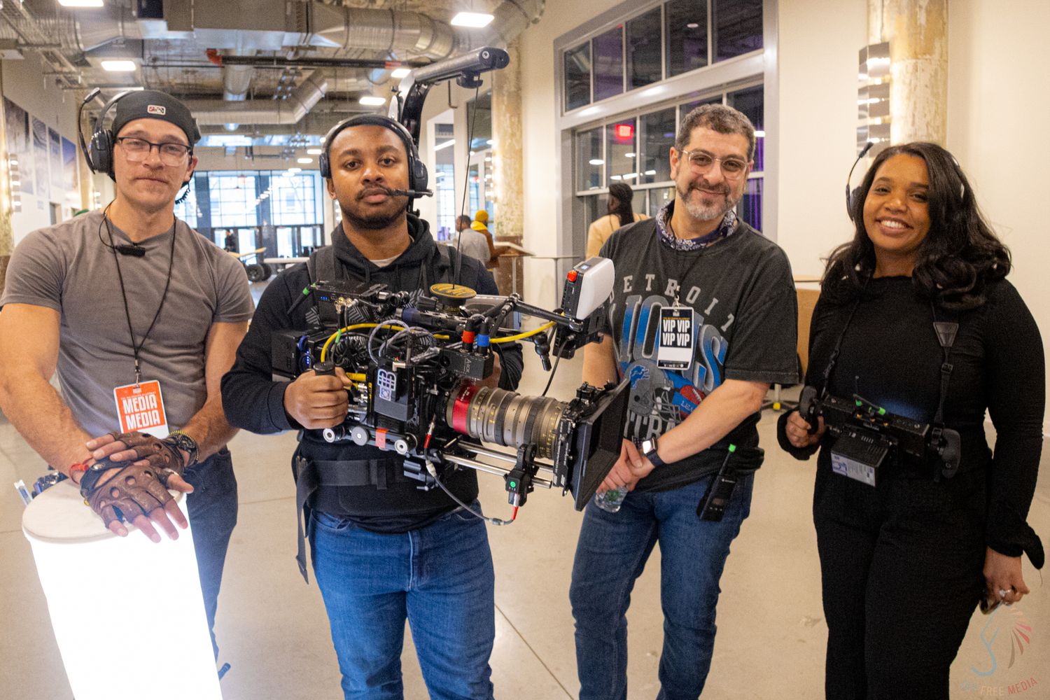 Four film crew members smiling while holding a professional camera rig in an indoor hallway setting.
