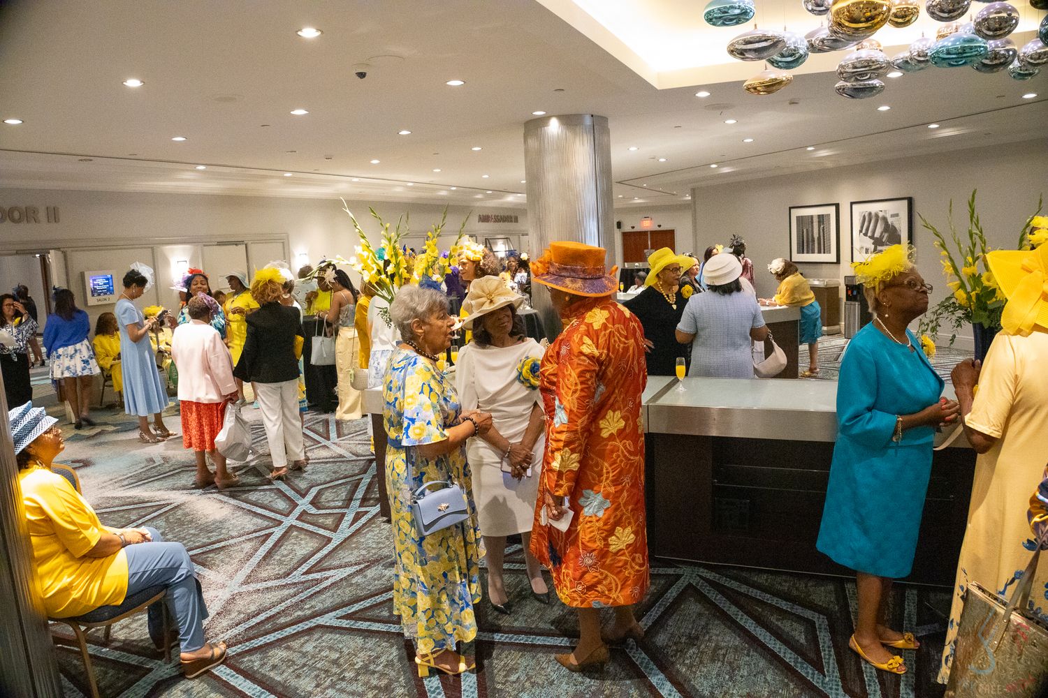Elegantly dressed guests in vibrant yellow and orange outfits mingle in a hotel lobby at a festive gathering.