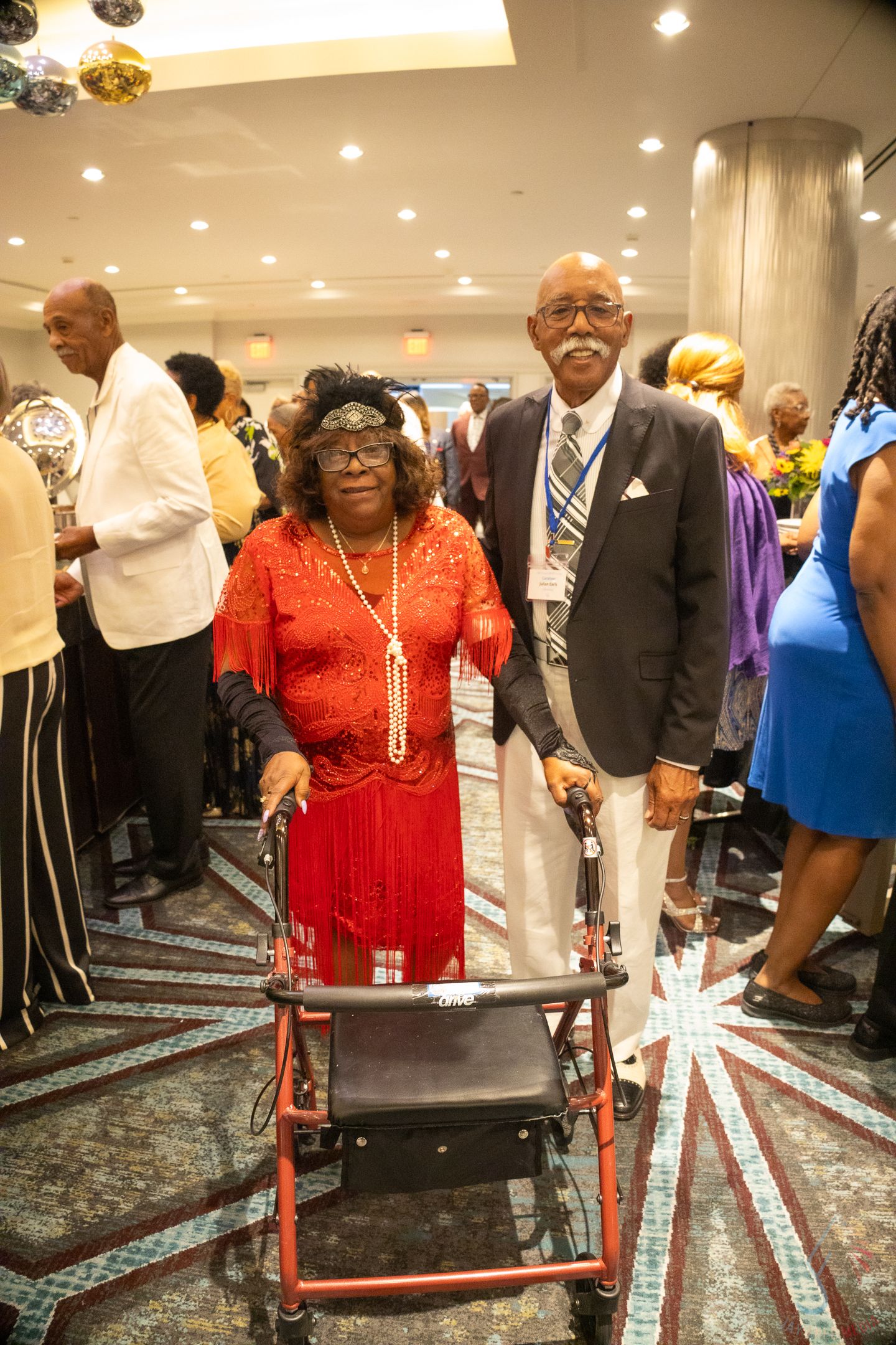 Elderly woman in red dress using a walker poses with a man in white pants at a formal event.