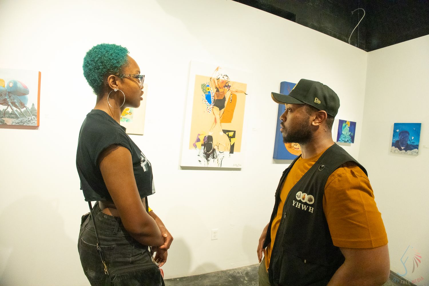 Two people conversing at an art gallery with colorful paintings displayed on white walls behind them.