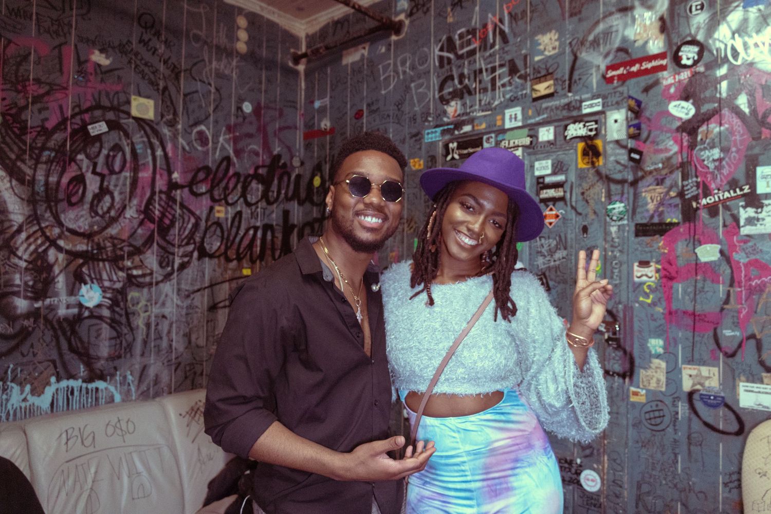 Two friends pose and smile in front of a graffiti wall, woman flashing peace sign wearing purple hat and holographic outfit.