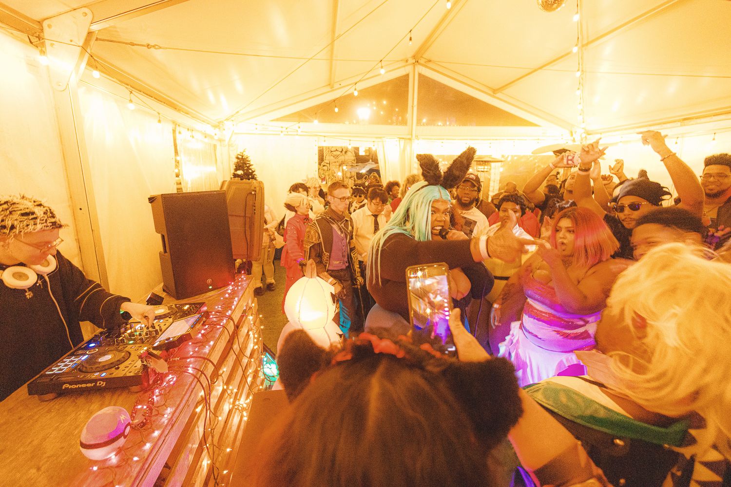 DJ performing at a lively festival party in a tent with colorful lights and costumed dancers.