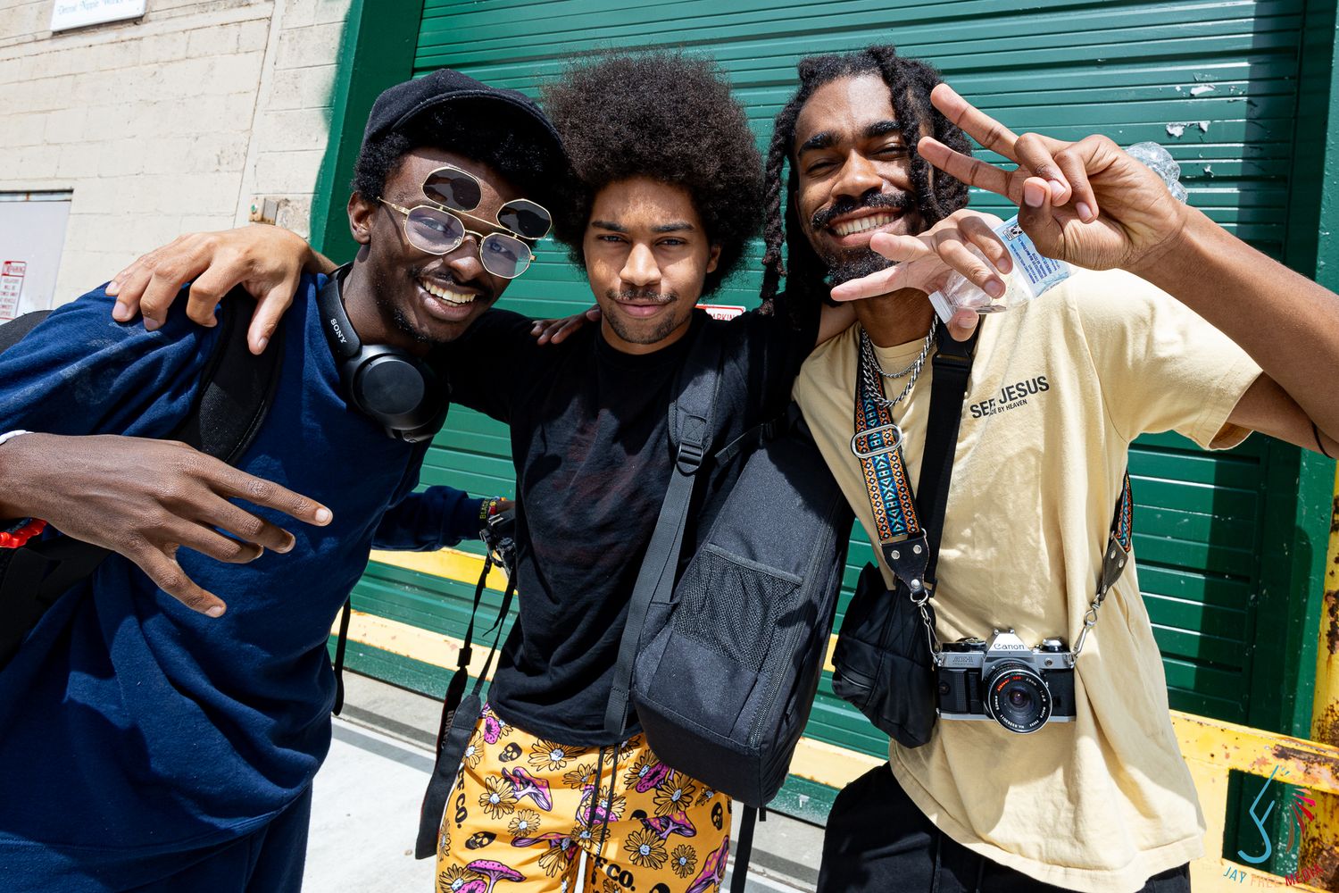 Three young men smiling and posing together outdoors, one making a peace sign, near a green wall.