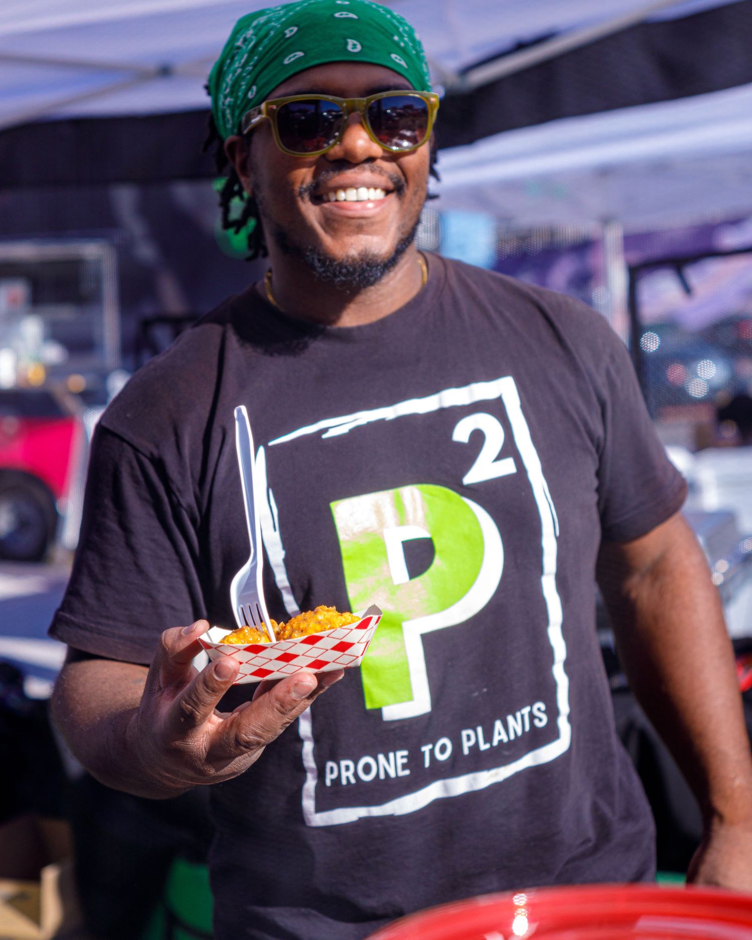 Smiling man wearing a Prone to Plants t-shirt and green bandana holds a plate of food at an outdoor event.